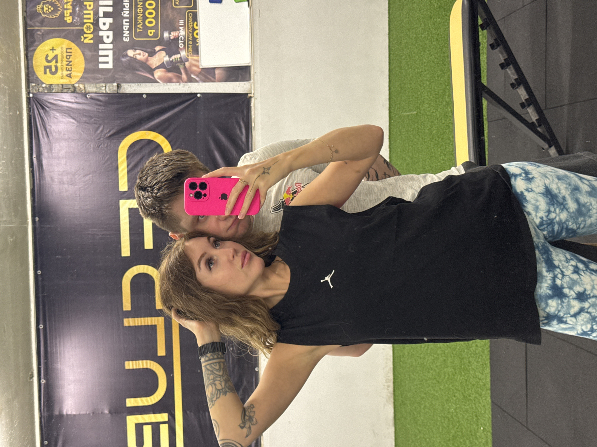 An indoor mirror selfie captures a man and woman, likely a couple, in what appears to be a gym or fitness center in Chaikovsky, Russia. The image is rotated 90 degrees counter-clockwise. The man, with short, light brown hair, is taking the photo, holding a vibrant pink iPhone with multiple cameras, partially obscuring his face. He wears a light-colored t-shirt. The woman is prominently featured in the foreground, looking slightly upwards with a neutral expression. She has wavy, light brown hair, and visible tattoos, including floral and abstract designs, on her left arm. She is wearing a black t-shirt with a white Jordan logo and distinctive blue and white tie-dye pants. A black wristband is on her left wrist, and she has a ring on her right hand.
The background, visible in the mirror reflection, includes gym elements: a white wall section, green artificial turf, and dark gray flooring or matting. On the right, a black metal structure with hooks, possibly for weights or resistance bands, is visible. Two large banners hang on the wall behind the subjects' reflection. The text on these banners is mirrored and fragmented due to the reflection. Partially visible and reversed words include "1000", "000 b", "+S2", "A", "CECINE", "ECTURE", "PMON", and "ILUPIM", suggesting the original text might be Cyrillic. The overall atmosphere suggests a relaxed moment, possibly after a workout.