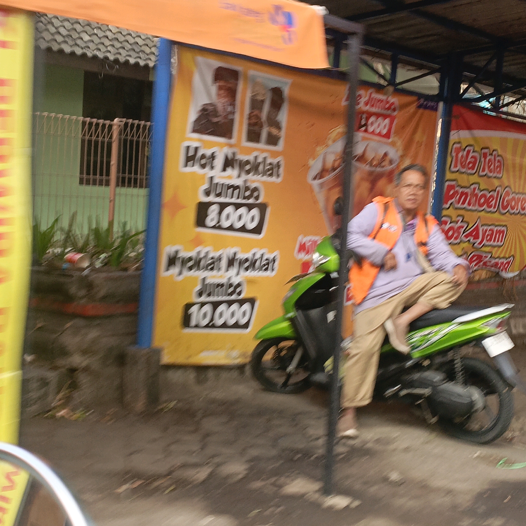 The image depicts a street scene in Salatiga, Indonesia, with a focus on a food stall and its proprietor. A man, wearing a light purple long-sleeved shirt and tan pants, is seated on a bright green motorcycle. He has an orange safety vest over his shirt. He is looking towards the camera, and his left leg is crossed over his right.
Behind the man and motorcycle are large, colorful banners advertising food products. One banner, predominantly orange, displays images of food items and text in Indonesian, including "Hot Nyeklat Jumbo" and prices such as "8.000" and "10.000". Another banner, to the right, is yellow with red lettering and appears to advertise "Ida Tela" and "Pinhoel Core" along with "Sofi Ayam". A third banner, a bright yellow, is partially visible on the far left.
The environment suggests an outdoor setting, possibly a roadside market or food court. The ground is paved with a rough texture, and there is some vegetation visible in planters. A metal fence with a simple pattern is seen behind some of the banners. The overall impression is a busy, everyday scene in a commercial area.