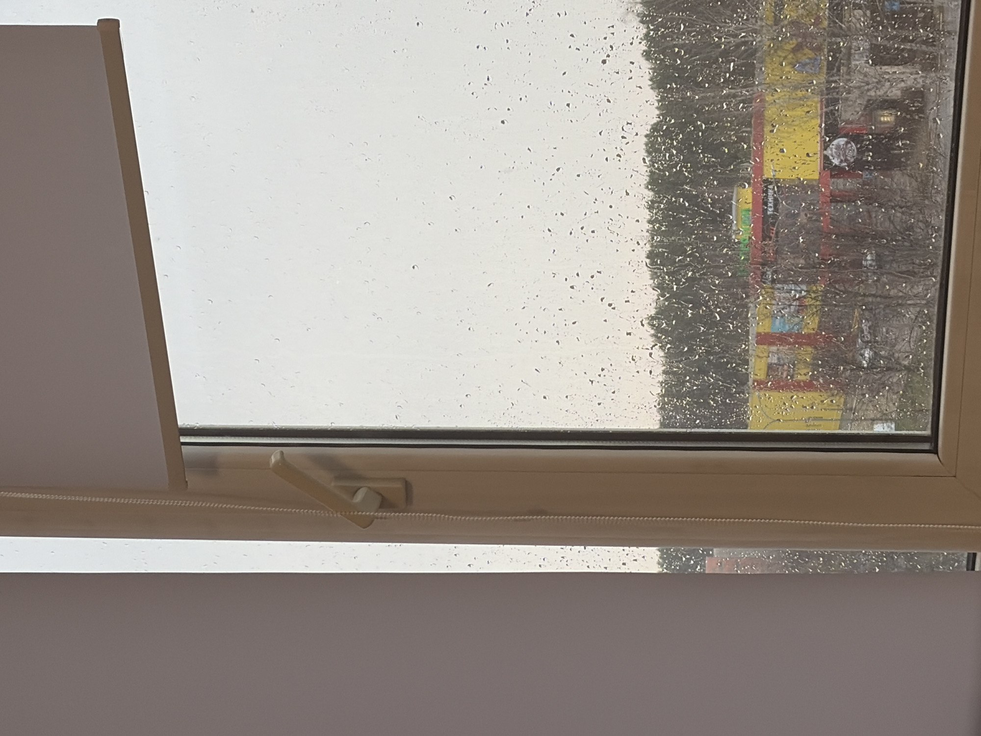 The image shows a view through a rain-streaked window. Outside, blurred by the rain, a building with a yellow facade and red structural elements is visible, along with trees lacking foliage. The window frame is light-colored. A portion of a white roller blind and its control mechanism is visible within the window frame. Based on the building exterior's color and structural appearance, the location is likely in Chaikovsky, Russia.