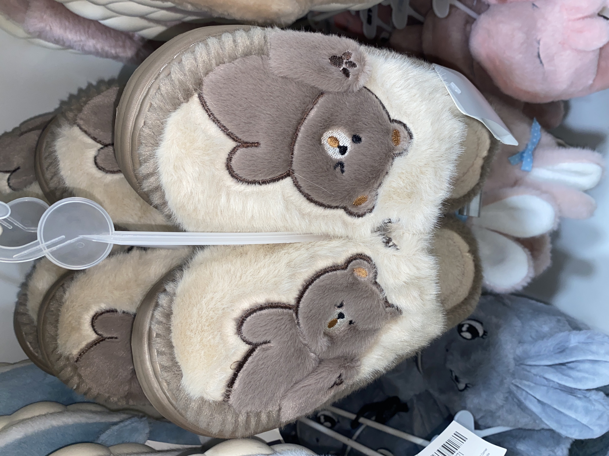 This close-up, vertically oriented image captures multiple pairs of plush slippers displayed in what appears to be a retail setting, likely in Davao City, Philippines. The primary subjects are two prominent pairs of slippers, stacked or hung, each featuring a large, embroidered teddy bear design on a creamy white faux fur top. The teddy bears are a soft brown or gray color, depicted lying down with embroidered faces including dark eyes, a small nose, and mouth, and lighter accents on the ears and muzzle. One bear also shows embroidered paw pads on its outstretched arm. The slippers have light brown or tan-colored soles visible along the edges.
In the background, other plush items are partially visible and out of focus, including pink-colored stuffed animals (one with a blue ribbon) and a gray plush toy with large ears, possibly a rabbit or elephant, towards the bottom right. Clear plastic display hangers or fasteners are seen holding the slippers. One white plastic tag, possibly a price tag or product information label, is attached to a slipper, displaying a barcode and partially visible text, including the numbers "771" and the word "Corner" or a similar fragment.
The scene suggests a merchandise display, highlighting soft, comfortable, and cute-themed footwear. The overall color palette is muted with creams, browns, grays, and soft pastels.