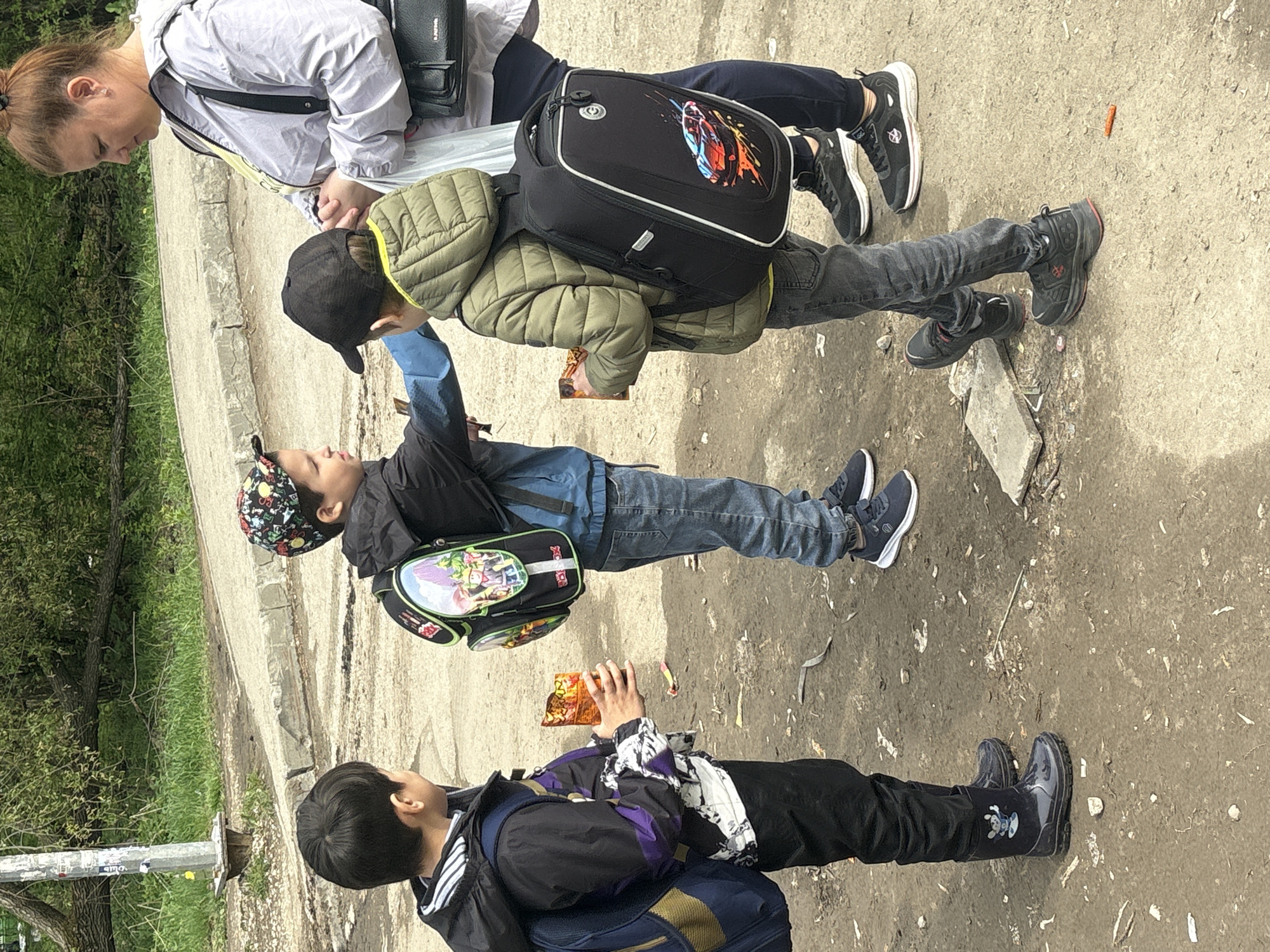 The image depicts a group of four individuals (one adult and three children) standing on a pavement adjacent to a road in Perm, Russia. The surface is gray concrete, with visible signs of wear and patches of dirt.
The adult, a female, is dressed in a light-colored jacket. She is looking down, and there are visible bags/purses at her shoulder and back.
The children are all wearing backpacks and casual clothing. The child closest to the adult wears an olive-colored jacket and a baseball cap. The next child wears a blue denim jacket, and a cap with a floral pattern. The last child is wearing a black jacket with white and purple stripes and a blue backpack.
The children appear to be eating from a pack of chips or snacks. The setting is a street with signs of urban development and nature, indicated by the presence of trees. The scene is captured in daylight, with a clear view of the subjects and their surroundings.