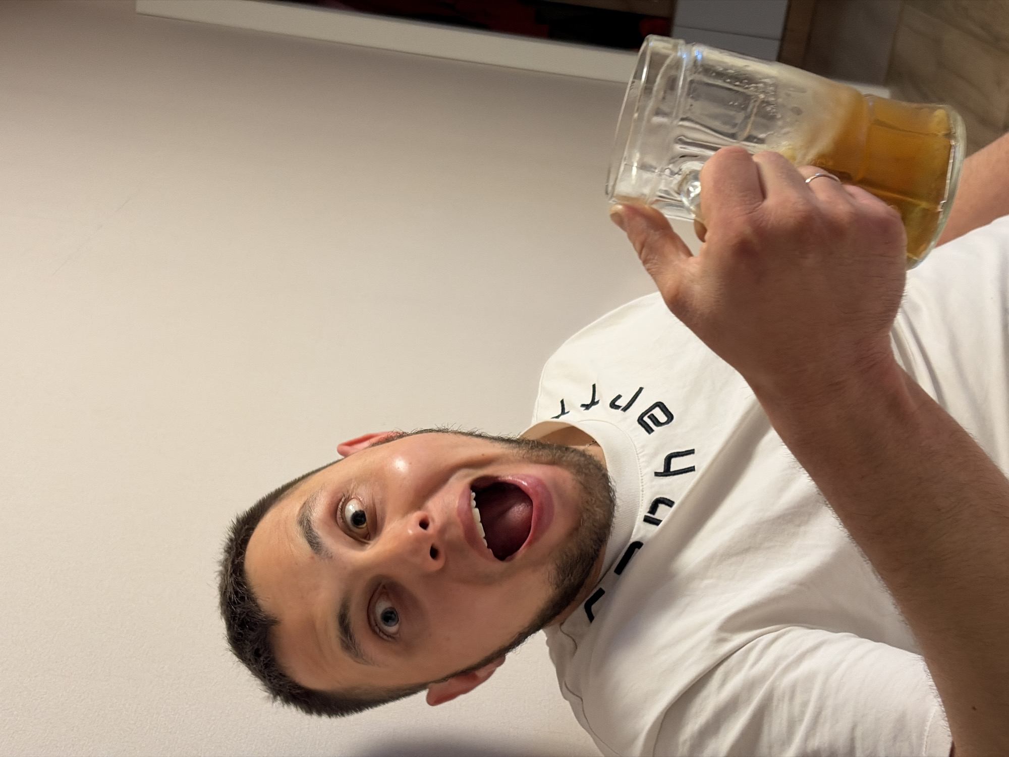 A young man with short dark hair and a beard is captured in a dynamic, rotated close-up, seemingly indoors against a plain light-colored wall in Krasnodar, Russia. He wears a white t-shirt with partially visible, stylized dark text, which appears mirrored or inverted, curving around the collar. The man holds a clear glass mug, containing an amber-colored liquid (possibly beer, cider, or iced tea), in his right hand. A simple silver-toned band ring is visible on his right ring finger. His face is turned towards the camera, mouth wide open and eyes wide, expressing an exaggerated emotion of surprise or excitement. A distinctive feature is his heterochromia, with one eye appearing blue/green and the other brown/hazel. The mug is tilted, suggesting movement or the act of drinking. The background includes a faint, blurred darker object at the top edge, likely a piece of furniture.