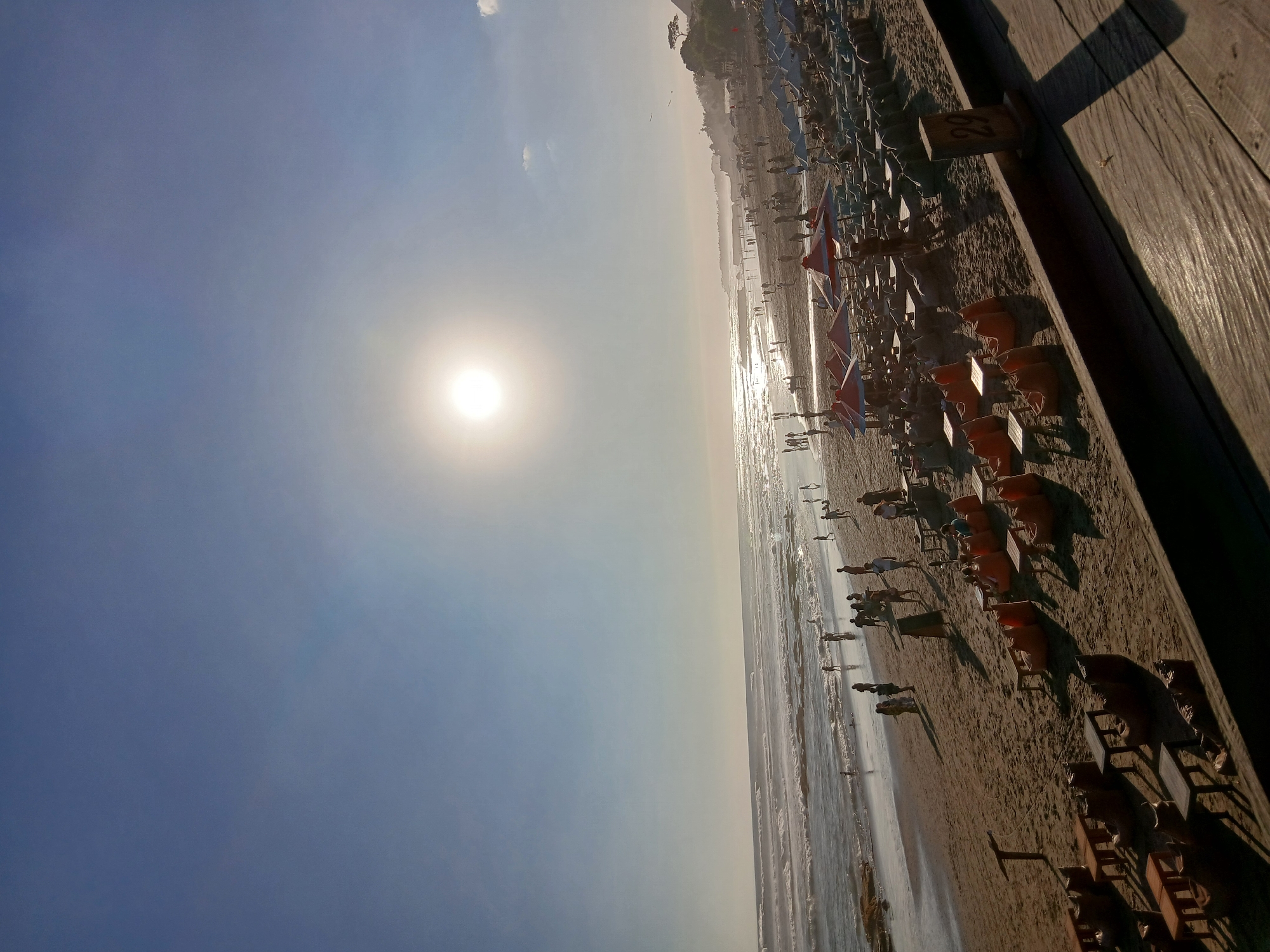 The image depicts a crowded beach scene during daylight hours, likely in North Kuta, Indonesia. The sun is high in the sky, casting a bright glare and creating reflections on the ocean. The water is moderately wavy, with several people visible wading and swimming near the shore. The beach itself is sandy and populated with numerous individuals, some walking, some sitting. In the foreground and midground, there are rows of lounge chairs and umbrellas set up for beachgoers, many of which are occupied. Scattered throughout the sand are tables and beanbag chairs, suggesting a casual seating area. A wooden walkway or deck is visible in the lower right corner of the image, casting a long shadow. In the background, a line of trees and some structures can be seen along the coastline. The overall atmosphere is one of a busy, popular beach destination.