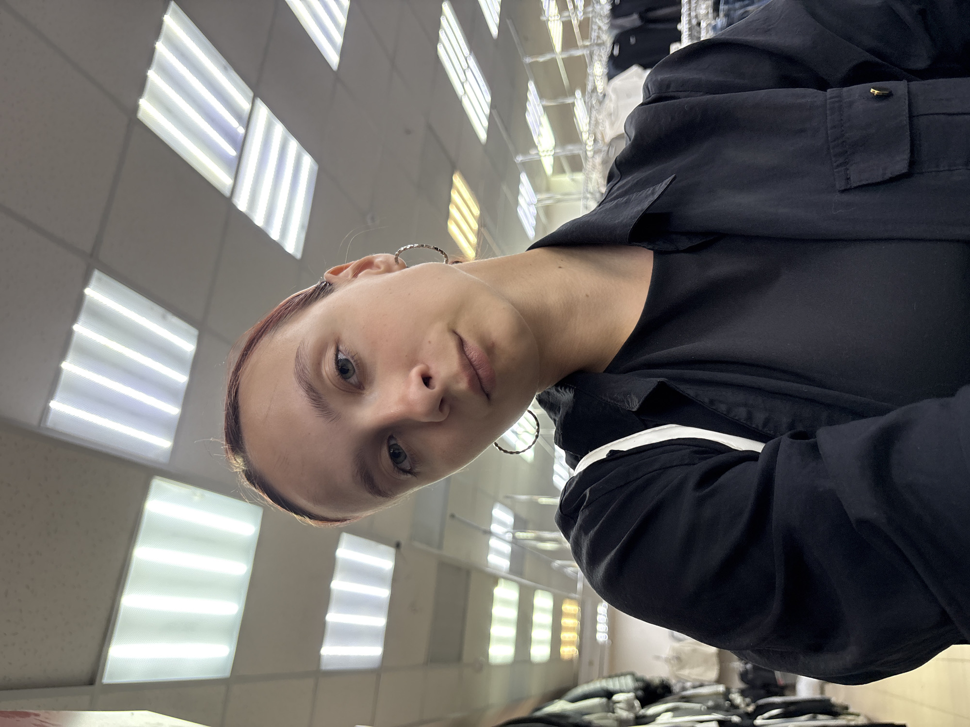 This rotated selfie features a young woman looking directly at the camera with a neutral expression. She has fair skin, light-colored eyes, and her hair, which appears to have a reddish-brown tint, is pulled back. She is wearing a black collared shirt or jacket over a dark-colored top and has small, metallic hoop earrings.
The background primarily shows a ceiling filled with rows of rectangular fluorescent light fixtures. Most of these lights emit a bright white glow, while some further back have a warmer, yellowish hue. The lighting suggests a well-lit indoor commercial environment. Blurred in the background, particularly on the right side, are racks that appear to hold clothing or other merchandise, reinforcing the impression of a retail store or department store.
The activity depicted is a self-portrait. The image is brightly lit by artificial overhead lights, making it impossible to determine the time of day or outdoor weather conditions. There is no visible text in the image. The setting is an indoor commercial space located in Votkinsk, Russia.