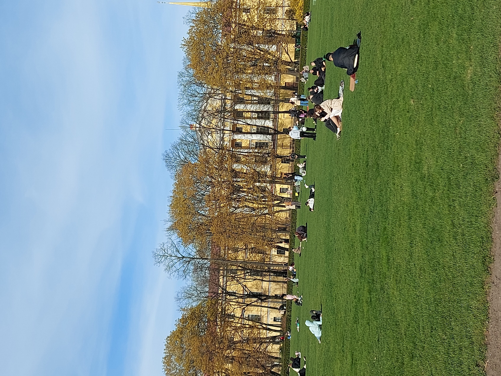 The image depicts a grassy lawn in Saint Petersburg, Russia, with people scattered across it. A large, light-yellow building with columns stands behind the lawn, framed by trees with budding leaves. The sky is blue with some light clouds. People are lounging, sitting, and standing on the grass.