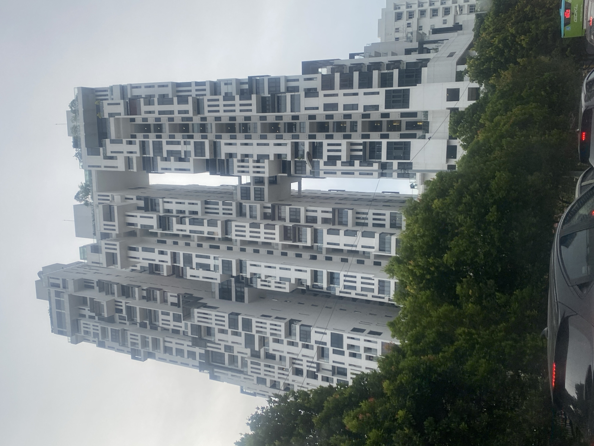 The image shows a modern apartment building in Kuala Lumpur, Malaysia, with a distinctive interconnected design. The building is tall and features a white facade with a pattern of dark windows and balconies, creating a visually striking and complex architectural style. A skybridge connects two sections of the building at a high level, with greenery visible on top of this connecting structure. The building is partially obscured by lush green trees in the foreground, suggesting it is located in an area with significant vegetation. In the lower part of the image, the rear of a black car is visible, indicating that the photo was likely taken from a street or road. The sky is overcast, contributing to a somewhat muted lighting condition. Another building, also white, is visible to the right, appearing to be part of the same development or a similar architectural style. A green bus with "GO KL" lettering is also visible in the lower right corner, suggesting the location is a public area with transportation services.
