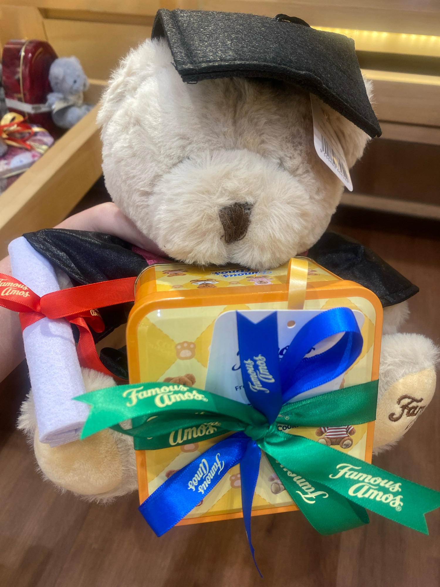 A close-up, eye-level shot captures a plush teddy bear, adorned with a graduation cap and gown, holding a scroll and a gift box. The teddy bear is positioned centrally in the frame. The gift box, a bright yellow tin featuring bear graphics and the "Famous Amos" logo, is tied with blue and green ribbons, each imprinted with the same brand name. A red ribbon is wrapped around the scroll, also displaying "Famous Amos." The background is blurred, showing wooden shelving and other retail items, suggesting an indoor commercial setting, possibly a gift shop or a section of a department store. The lighting appears to be artificial, casting a warm glow on the subjects. The overall scene is festive and celebratory, likely related to graduation or a gift-giving occasion. There is no clear indication of the location within Kuala Lumpur, Malaysia, beyond it being an indoor commercial space.