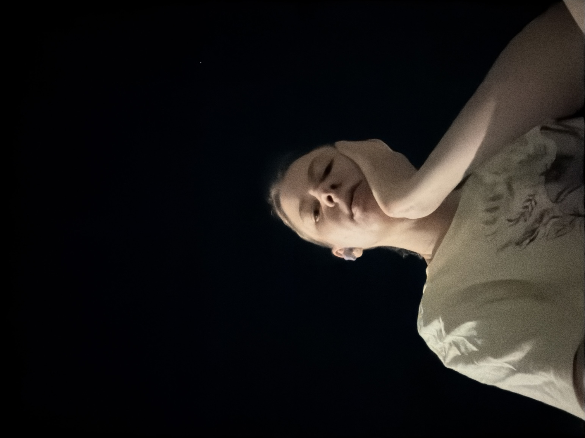 The media file depicts a person's upper body from a low-angle, wide shot against a dark background. The person is positioned in the right half of the frame, with their head tilted and their right arm raised, hand resting on their head. They are wearing a light-colored t-shirt with a subtle pattern, possibly floral or abstract, on the chest. A small, faint light source is visible in the upper left quadrant of the frame, suggesting a distant light or star. The scene is dark, implying it is nighttime or a very dimly lit indoor space. The orientation of the camera and the cropping suggest the image might be a selfie or a candid shot taken from a low vantage point. The overall mood is casual and somewhat introspective, with the person's pose conveying a relaxed or thoughtful state. No specific text is visible in the image. Given the location context of Dongducheon-si, South Korea, the setting could be outdoors at night, perhaps a park or a residential area, or an indoor environment with minimal lighting.