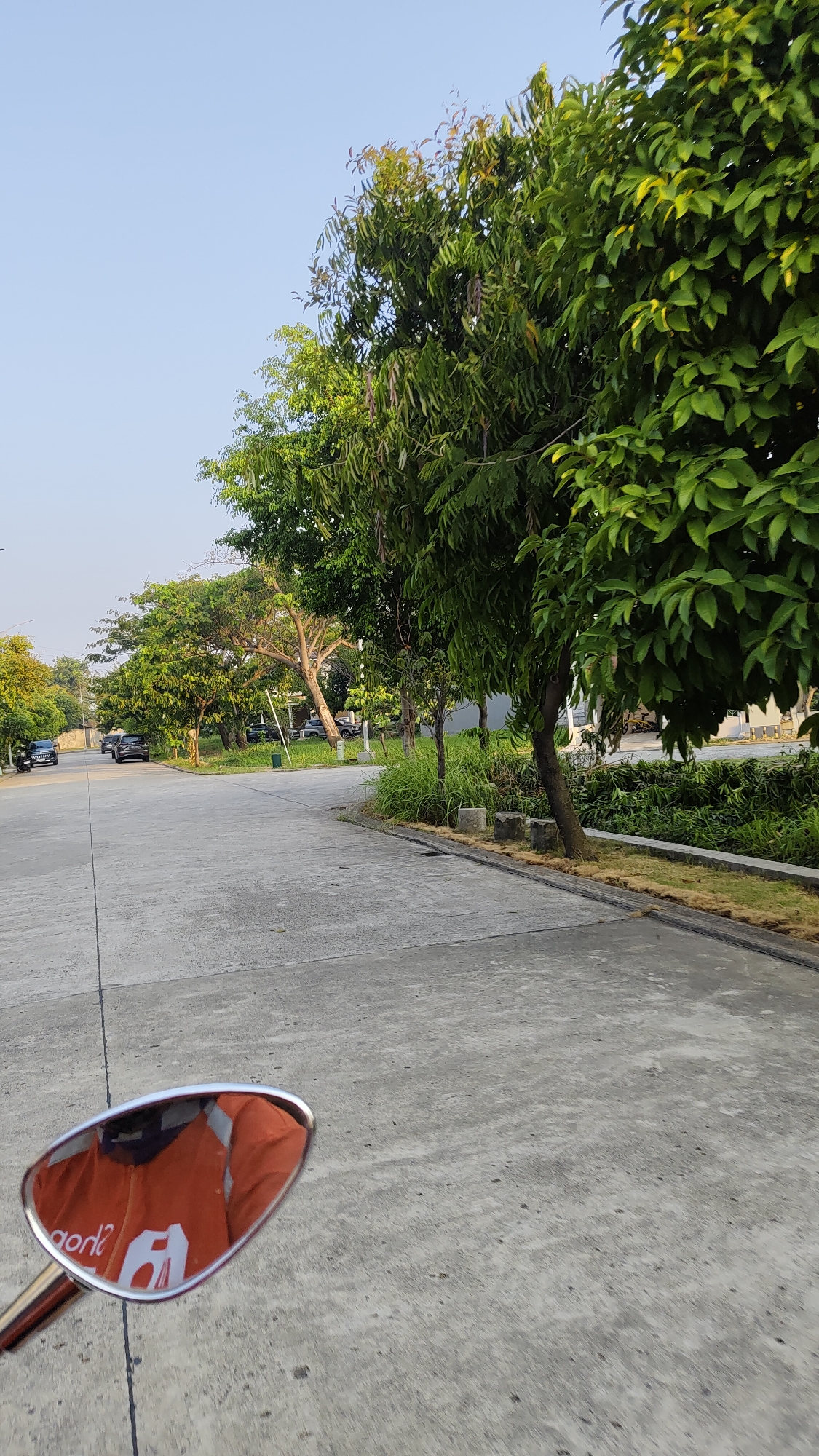 The image depicts a paved road in a residential area, possibly in Kebalen, Indonesia, lined with trees and vegetation. A motorcycle's side mirror in the foreground reflects an orange jacket with white lettering. In the background, several cars are parked along the road. The sky is clear and blue, suggesting daytime. The overall scene is serene and orderly.