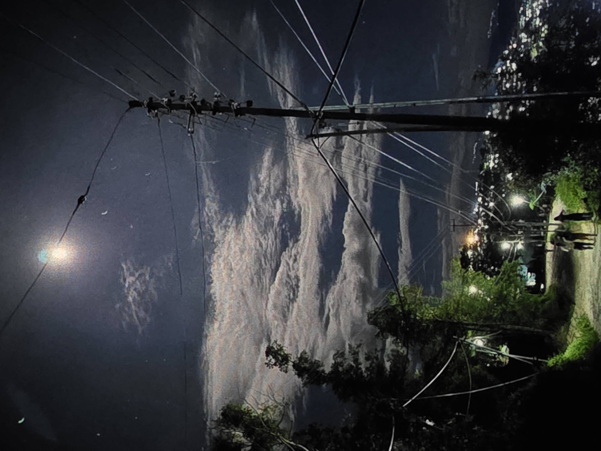 The image depicts a night scene with a full moon illuminating the sky, casting a soft glow on the clouds. A paved road winds through a rural or semi-urban area, with trees and vegetation lining its sides. Several power lines stretch across the frame, supported by utility poles, with distant city lights visible in the background, suggesting a settlement. A few individuals are walking along the road, their figures silhouetted against the faint light. The overall atmosphere is serene and somewhat mysterious, characteristic of a quiet night. There is no specific indication of the location being the city of Dubno, Ukraine.