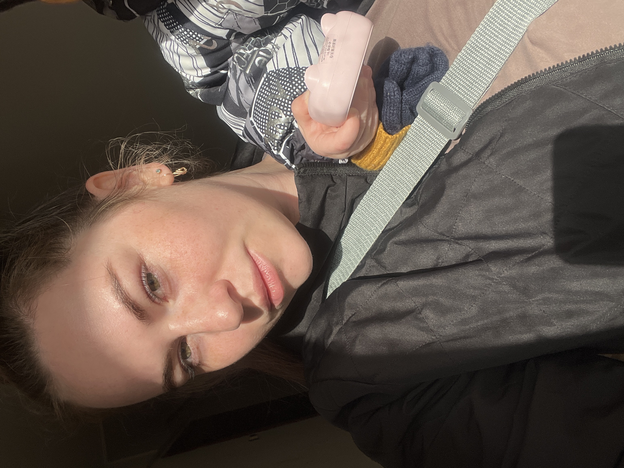 An adult female is depicted in a close-up, illuminated by direct sunlight from the right, creating strong contrast and shadows on her left side. Her hair is pulled back, revealing gold-toned hoop earrings and a stud in her right ear. She has green-hazel eyes and visible freckles on her cheeks. She wears a dark, possibly quilted jacket. A light grey woven strap with a plastic buckle crosses her upper body, suggesting she is wearing a carrier or seatbelt, potentially in a vehicle. To her right, a child is partially visible, clad in a black, white, and grey patterned hooded jacket, with yellow and dark blue knitted clothing peeking from beneath the sleeve. The woman holds a small, pink, cat-ear-shaped object in her right hand, on which faint text, possibly Chinese characters, is discernible. The background is dark and out of focus, consistent with an interior setting, likely a vehicle, in the city of Chaikovsky, Russia.