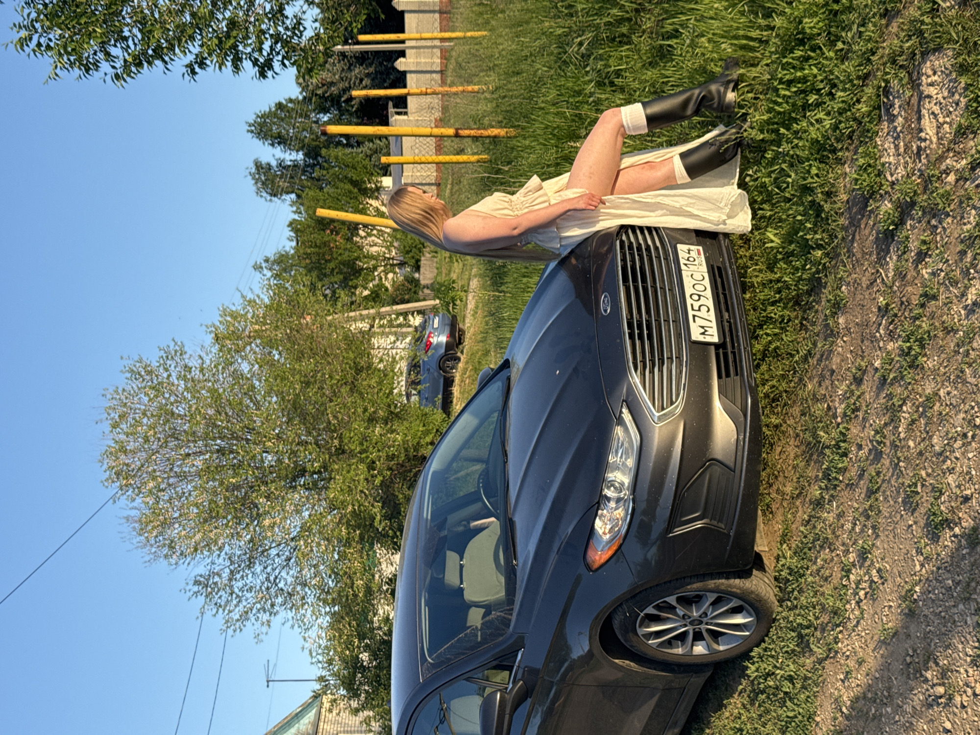 A woman in a light-colored dress and black boots is reclining on the hood of a dark gray Ford sedan. The car's license plate reads "M759OC164 RUS". The vehicle is parked on a grassy shoulder adjacent to a dirt or gravel road in what appears to be a rural or suburban area of Усть-Курдюм, Russia. A row of yellow metal posts runs along a grassy embankment in the background. A tree and sections of houses are visible in the background under a clear blue sky.