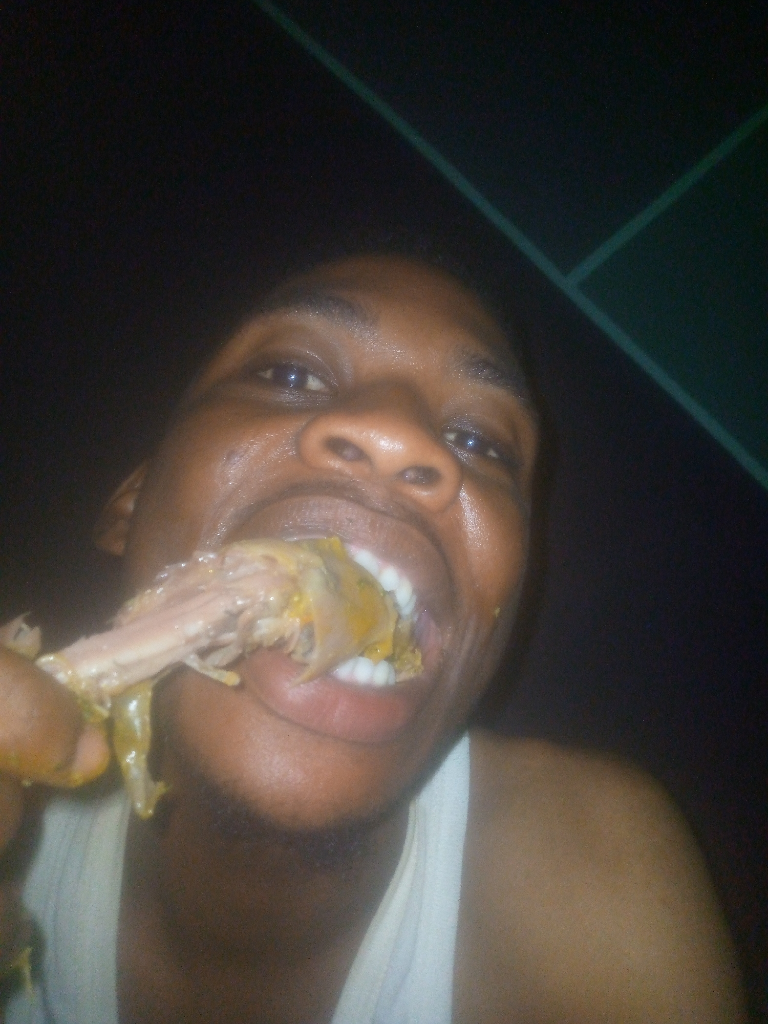 An adult male with dark skin is eating a piece of meat off the bone. He has the meat in his mouth. The background is dark, but there is a green geometric shape in the upper right corner. The man is wearing a white tank top. The location is not discernable from the image. The city of Ikorodu, Nigeria cannot be confirmed based on the image provided.