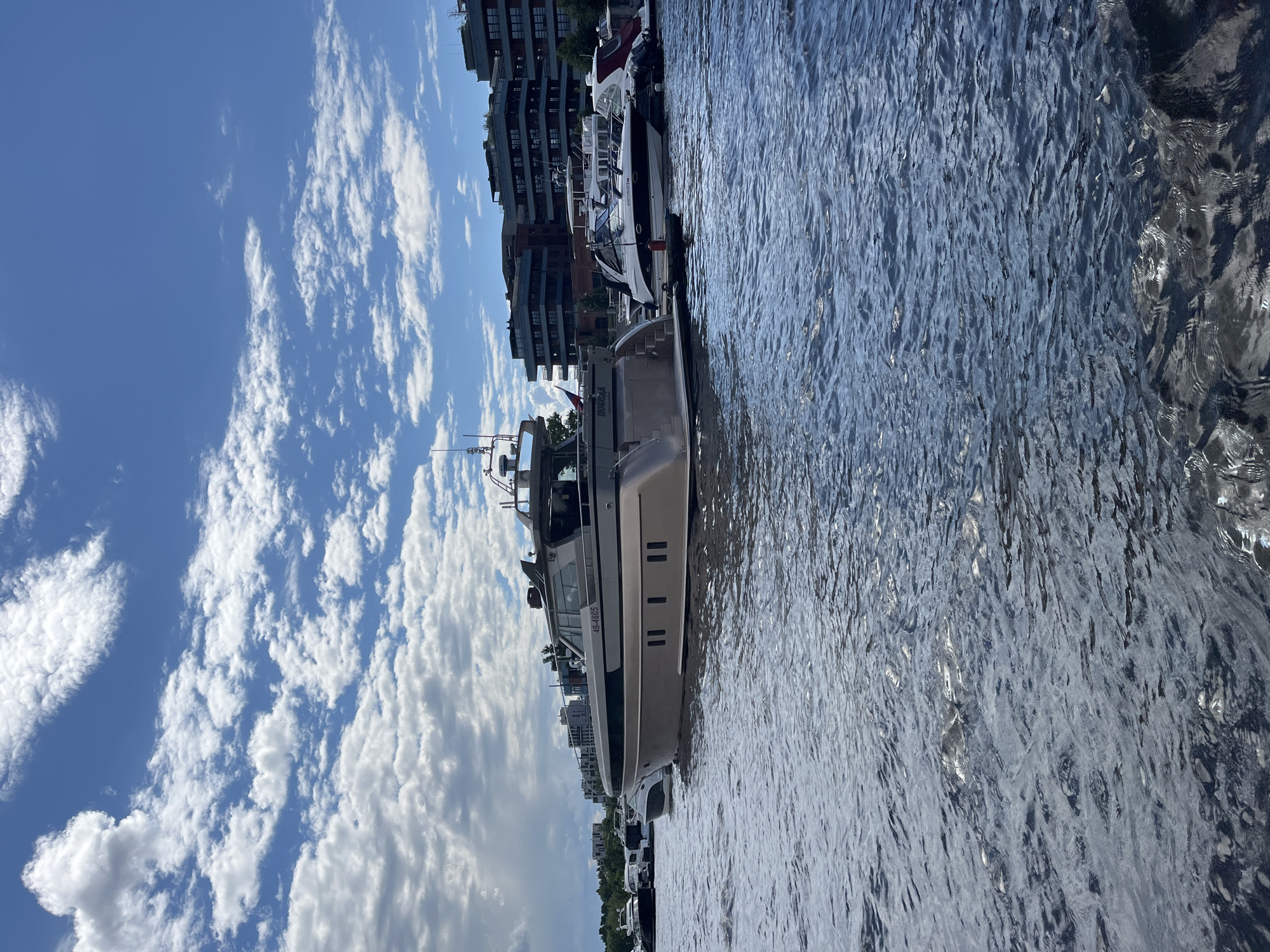 The image depicts a marina in Saint Petersburg, Russia. The foreground is dominated by rippling blue water, reflecting the sky and the surrounding structures. Moored along the embankment are several boats and yachts, with a prominent beige yacht in the center of the frame. This yacht has a sleek design with multiple windows and appears to be a luxury vessel. In the background, a modern, multi-story building with a brick facade lines the waterfront, suggesting an urban or commercial setting. The sky above is a vibrant blue, dotted with fluffy white clouds, indicating fair weather. The image is oriented vertically, with the sky occupying the left side and the water and boats on the right. A flag is visible on the beige yacht. The overall scene suggests a pleasant day at the marina, with boats docked and the city skyline visible. The number "49-4605" is visible on the side of the beige yacht. The lighting suggests it is daytime, likely late morning or afternoon, given the bright sky and shadows.