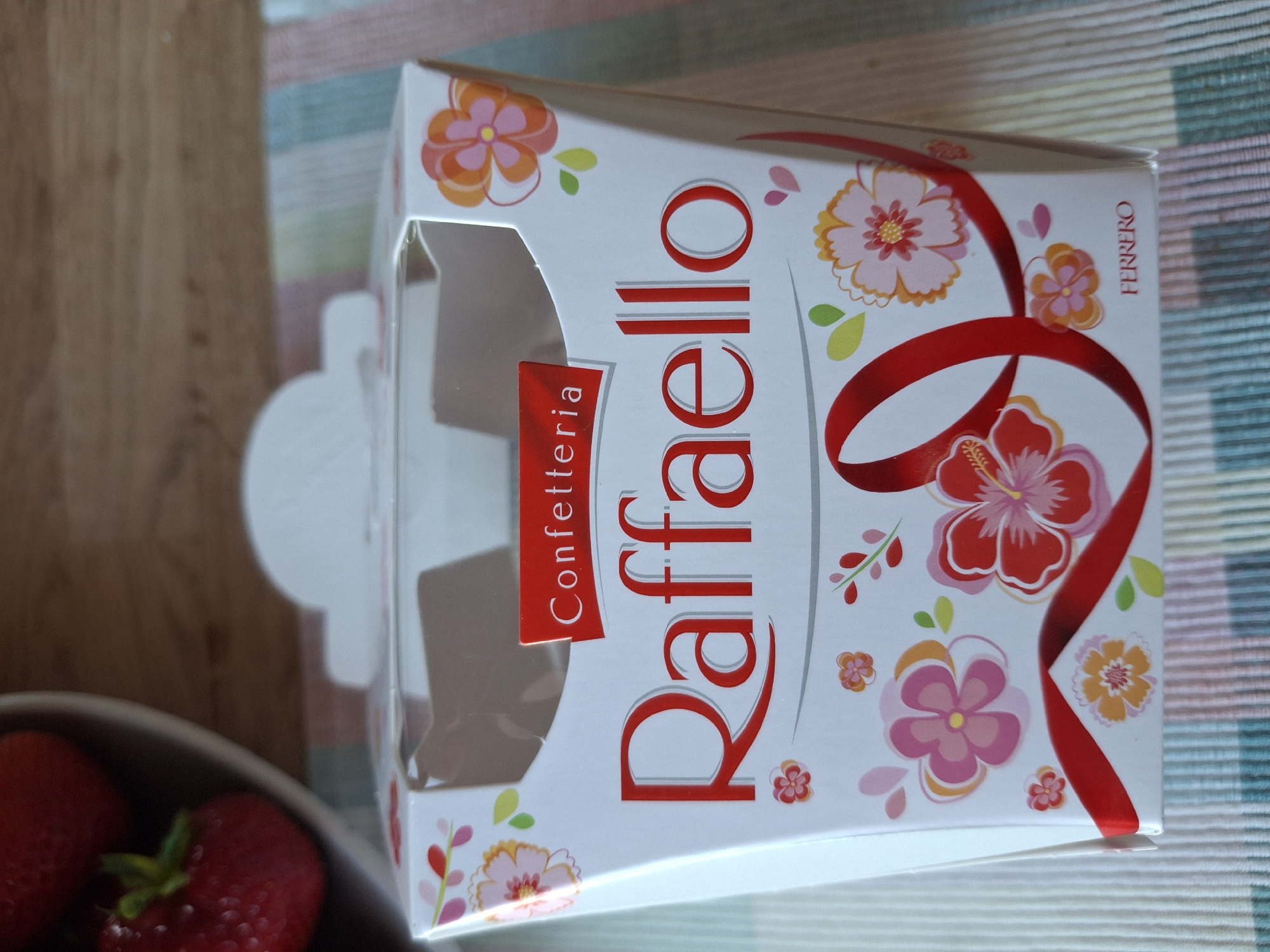 The image shows a close-up of a white and red Raffaello confectionary box with floral decorations and a red ribbon. The box is partially open, revealing a clear plastic insert. The text "Confetteria Raffaello" is prominently displayed on the front. The brand name "FERRERO" is visible at the bottom right of the box. In the background, a bowl of strawberries is partially visible on the left, and a striped tablecloth is underneath the box. The image appears to be taken indoors, with natural lighting. No specific location in Åsgårdstrand, Norway can be identified from the image content.