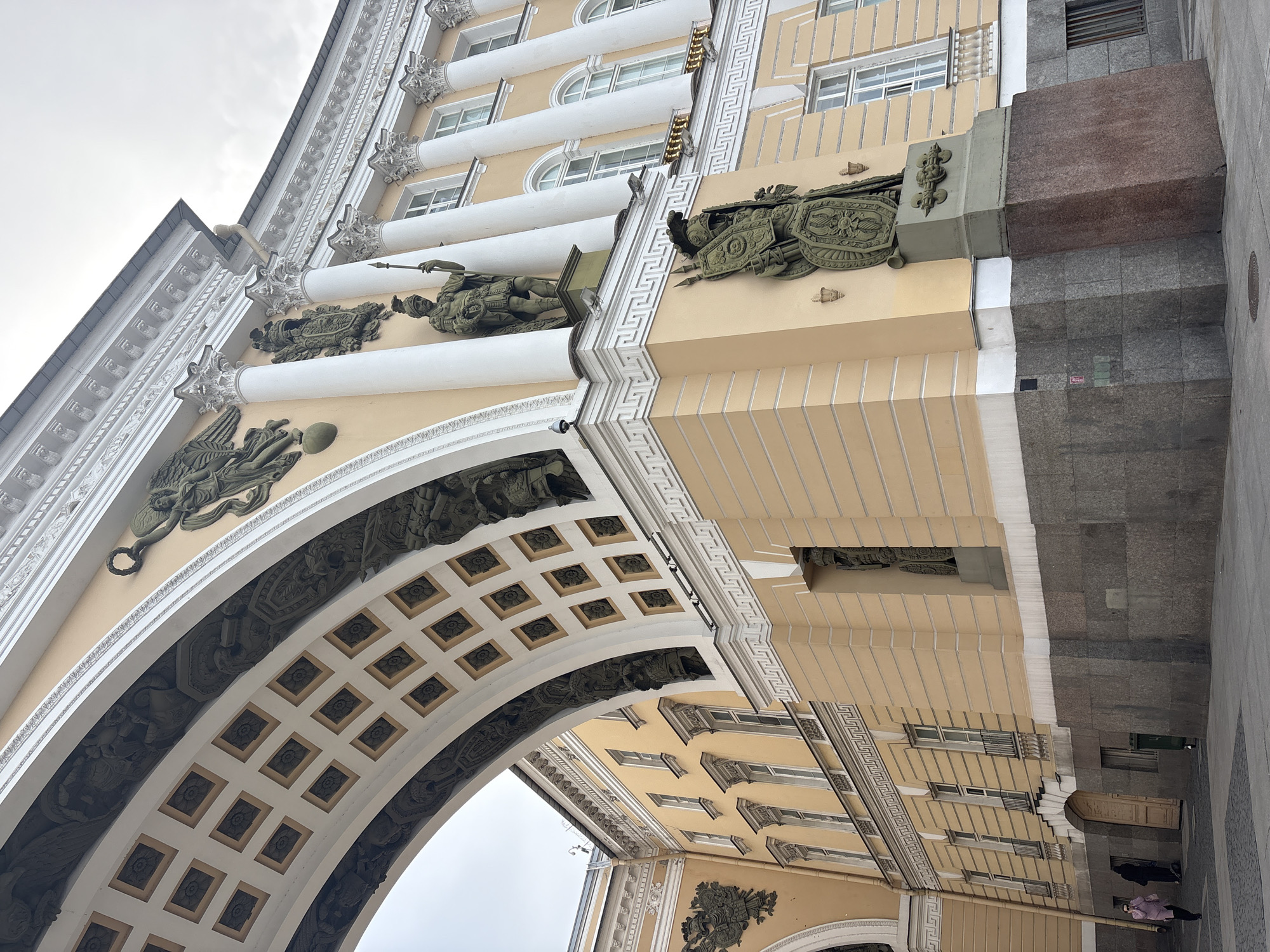 The image shows a low-angle view of the General Staff Building in Saint Petersburg, Russia. The architecture is neoclassical, featuring a large archway with coffered ceilings and ornate carvings. The building is primarily yellow, adorned with white columns and decorative elements. Bronze sculptures are positioned along the façade.
On the right side of the image, a section of the building transitions to gray granite, leading to the ground where pedestrians are walking.