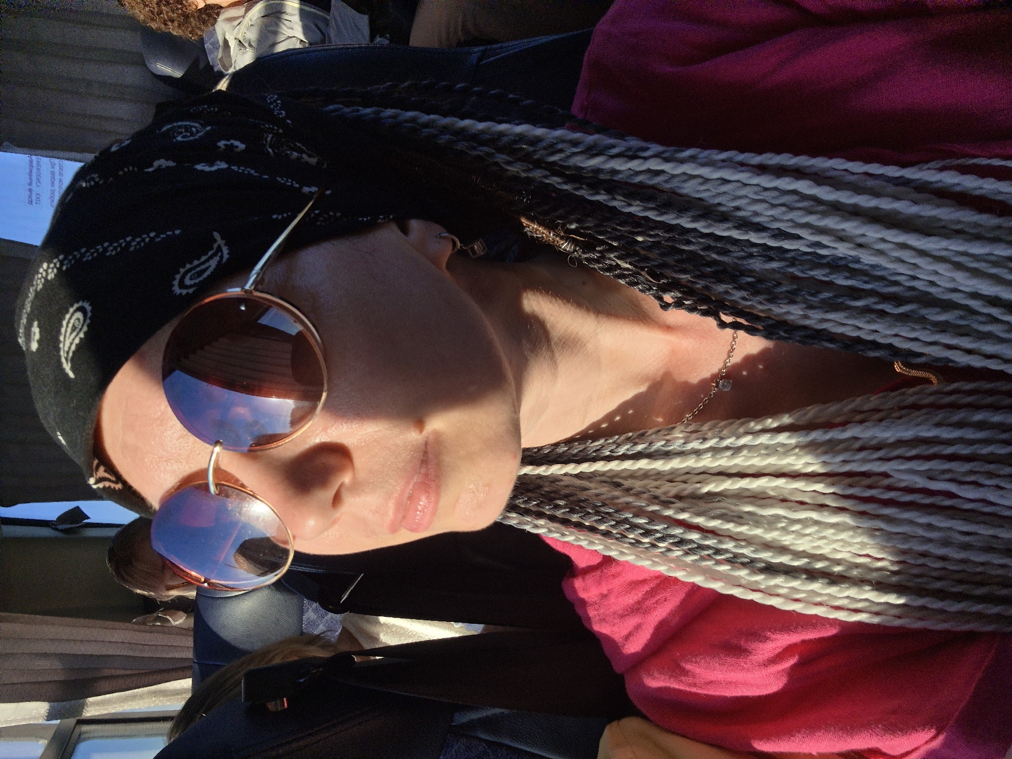 This is a close-up, waist-up, indoor shot of a woman with braided hair and sunglasses, likely taken on a bus. The woman is wearing a pink shirt and a black bandana. Sunlight is visible on her face. She appears to be looking to her left, away from the camera. In the background, there are blurred figures of other passengers and the interior of a bus. The exact location of Verkhniye Tatyshly, Russia, is not discernible from the image itself, but it is presented as the context for this photograph.