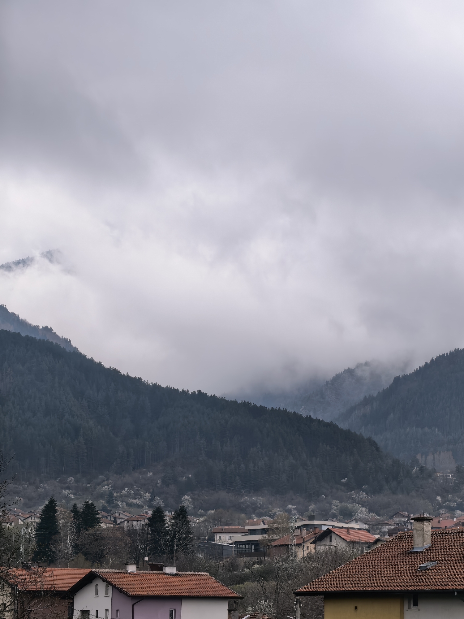 An overcast day in what appears to be a small settlement nestled in a valley. The low clouds and fog obscure the upper portions of the forested mountains that flank the settlement. In the foreground, the roofs of houses with red tiles dominate the view. Some houses are painted in shades of white, pink, and yellow. The scene is still and quiet.