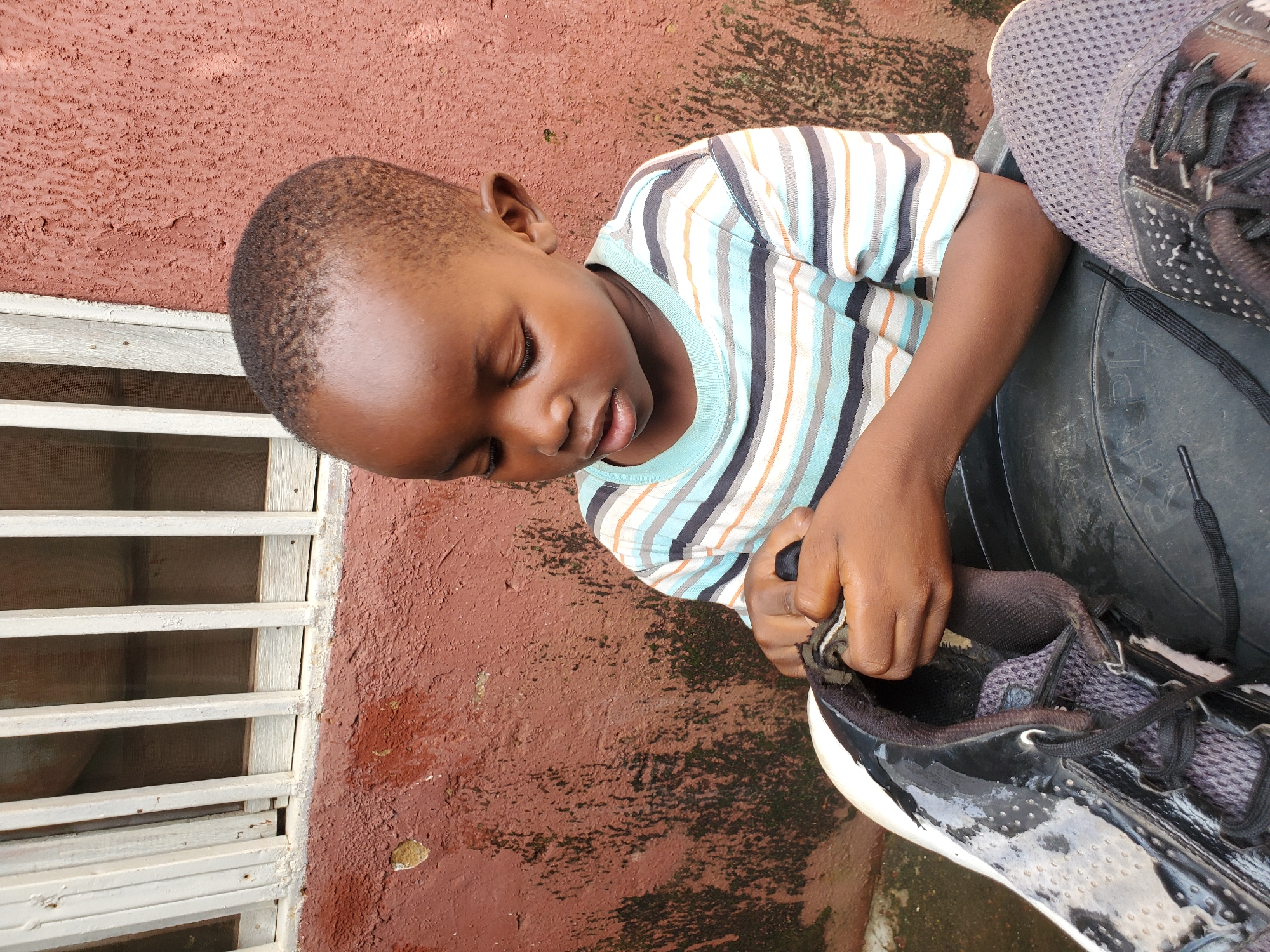 A young boy with dark skin and short hair, wearing a striped t-shirt, sits outdoors and appears to be examining a black and gray sneaker. He holds the shoe in his hands, focusing his attention on it. The background consists of a rough, reddish-brown wall with some staining and a window with white bars. The scene is captured in a close-up shot.