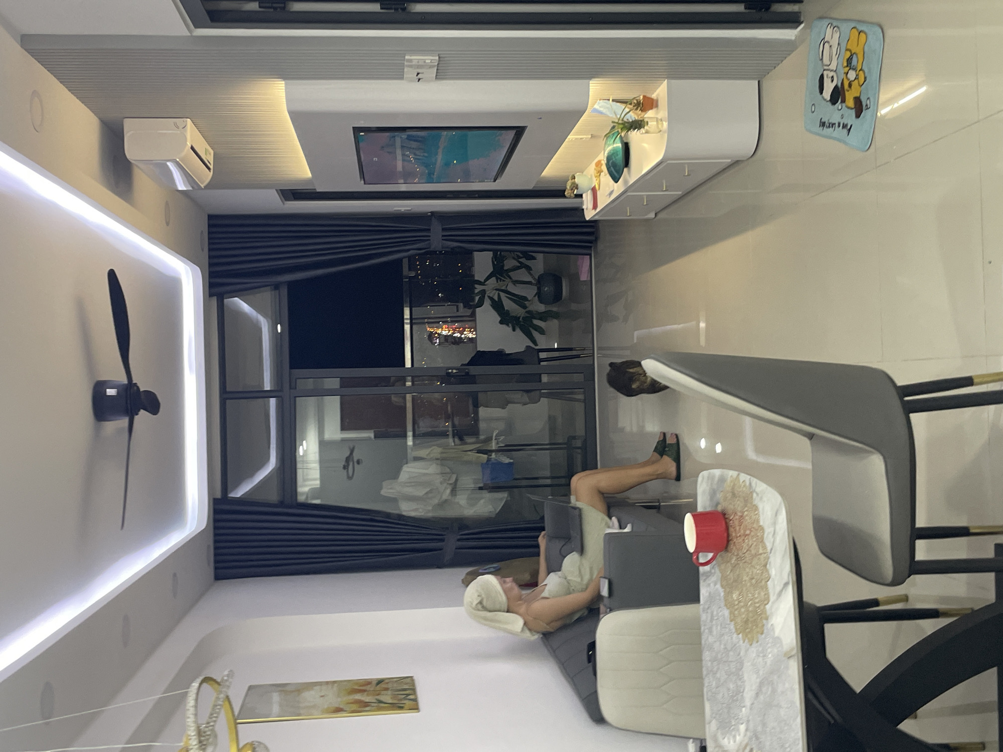 The image depicts an interior living room scene in Nha Trang, Vietnam. A woman with a towel wrapped around her head is seated on a dark grey couch, engrossed in using a laptop. She is wearing a light green dress and green sandals. The couch is positioned in a room with a tiled floor and a modern ceiling with embedded lighting. To the left of the couch, a dining table with a marble-like top and dark legs is set with grey chairs. A red mug sits on a decorative placemat on the dining table. Further into the room, a sleek white entertainment unit is mounted on a wall, featuring a large television screen displaying a landscape image. A white air conditioning unit is mounted on the wall above the entertainment unit. The room also includes several potted plants, and a small rug with cartoon dogs is visible on the floor. A large sliding glass door, partially obscured by dark curtains, offers a view of a balcony and the city lights at night. A black ceiling fan is visible on the ceiling, and a chandelier with gold accents hangs near the top left of the image.