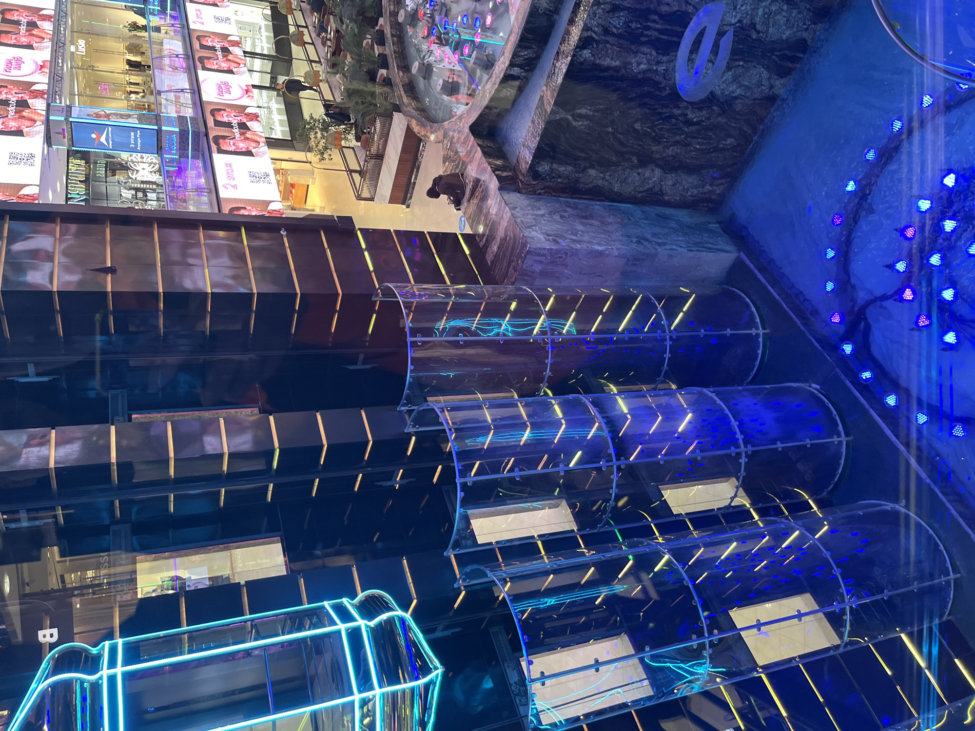 The image shows the interior of the Central Children's Store on Lubyanka Square in Moscow, Russia. The viewpoint is directed upwards, capturing the store's multi-level structure. Transparent, cylindrical tubes are arranged in tiers, adorned with blue and yellow lighting, creating a futuristic effect. At the top of the frame, elements of the store's upper levels are visible, including what appears to be a large decorative water feature and sections displaying advertising. The architectural features and lighting design dominate the scene.