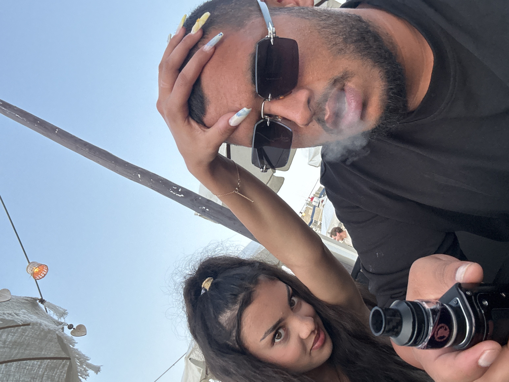 The media file is a selfie taken outdoors, likely during the day given the bright sky and illumination. The foreground features two individuals. On the right, a man with dark hair and a beard is visible, wearing a black t-shirt and sunglasses. He is holding a vape device in his right hand and appears to be exhaling smoke. His hand is positioned near his head, which is tilted back slightly, and he has long, light-colored fingernails with some nail art.
To the left and slightly below the man, a woman with long, dark, wavy hair is looking towards the camera. She is wearing a bracelet and has a playful or curious expression. Her arm is raised, and her hand is near the man's head.
The background shows a clear blue sky and some outdoor elements, possibly a beach or an outdoor seating area with umbrellas or canopies made of light-colored fabric, decorated with tassels and heart-shaped charms. There is also a thin pole with a small, woven lamp hanging from it. In the further background, another person is visible, wearing a light-colored shirt, suggesting a public or social setting.
The overall mood appears casual and relaxed, with the subjects engaged in vaping and taking a selfie. There is no visible text in the media file. The lighting suggests it is daytime, with no clear indicators of specific weather conditions other than a clear sky. The setting evokes a vacation or leisure atmosphere.