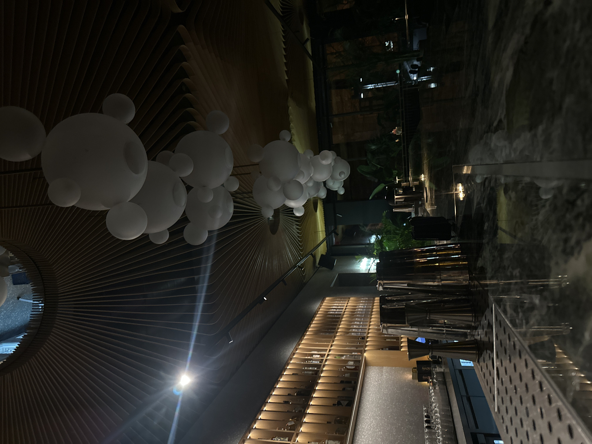 The image depicts the interior of a bar or restaurant, likely at night, with a view toward an outdoor patio area. A unique ceiling design features radial wooden slats and clusters of large, white, spherical decorations. Below the ceiling, a long shelving unit displays numerous bottles, presumably alcohol. In the foreground, a bar counter is visible, lined with metal bar tools. Through the glass wall, an outdoor seating area with greenery is visible. The location is potentially in Varna, Bulgaria, but there is no confirmatory evidence within the image itself. No people are clearly visible.