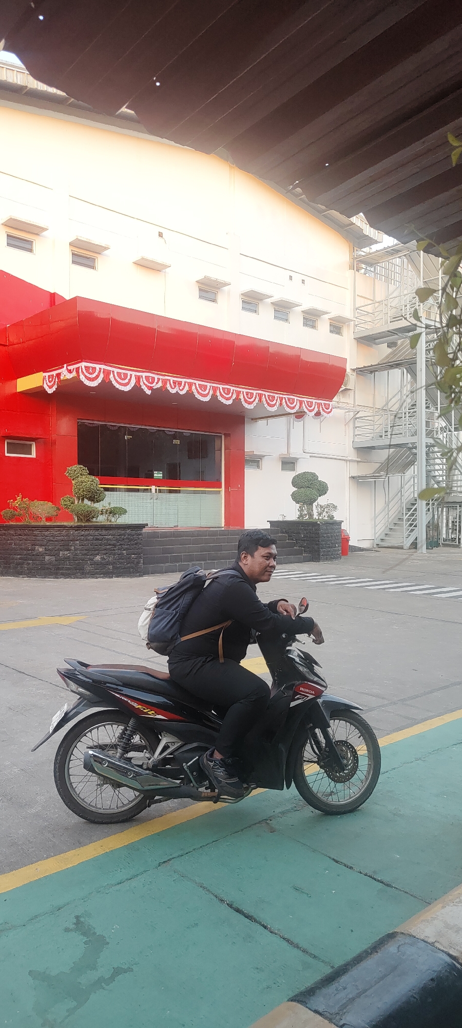 A man is sitting on a black motorcycle, his body angled to the right, and his head turned to the left. He is wearing a black long-sleeved shirt, black pants, and dark sneakers. A blue backpack is slung over his shoulders, with tan straps visible. The motorcycle has red and white decals. The man's left arm is resting on the handlebars.
The background features a modern building with a red facade and white accents. Decorative white and red pennant banners hang above the entrance, which has large glass doors. To the right of the building, a metal staircase is visible. In front of the building, there are two small, sculpted bushes in dark planters. The ground is paved with concrete and marked with yellow and green lines. The scene is lit by daylight, with a shadow cast by the man and motorcycle. The overall impression is an urban or industrial environment, potentially in Nusa Indah, Indonesia, given the architectural style and the presence of a motorcycle.