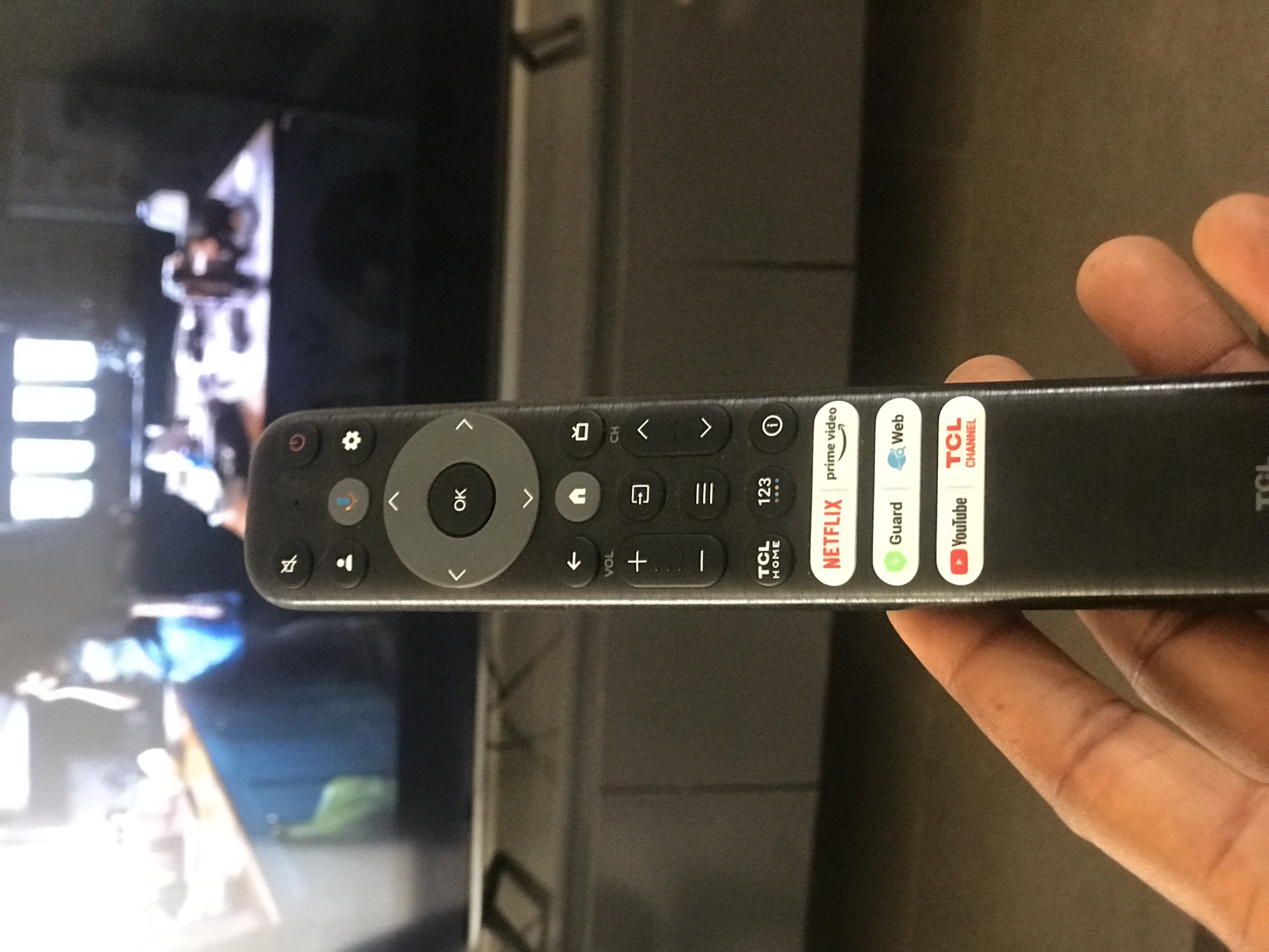 A person is holding a TCL brand TV remote control in front of a TCL television. The remote has buttons for Netflix, Prime Video, YouTube, and other streaming services. The TV screen shows an image of people. The background appears to be a living room with a television stand.