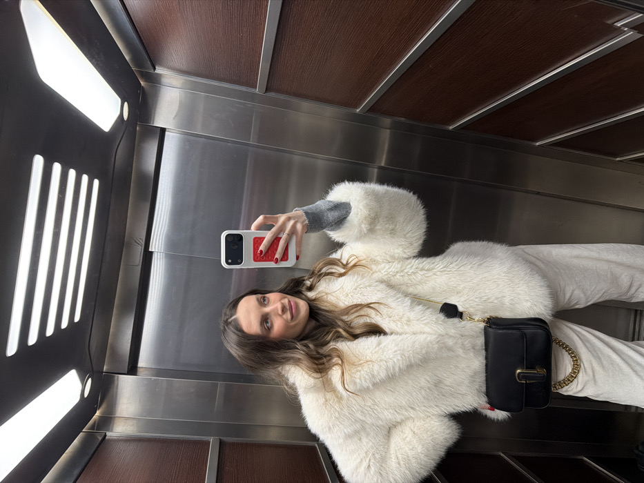 A woman poses in an elevator, holding a phone with a red and white case in her right hand to take a selfie. She wears a fluffy, off-white coat over a gray long-sleeved shirt and light-colored pants. A small, black purse with a gold chain strap hangs on her left side. The elevator features a metallic wall and dark wood panels above. Fluorescent lights are visible on the left.