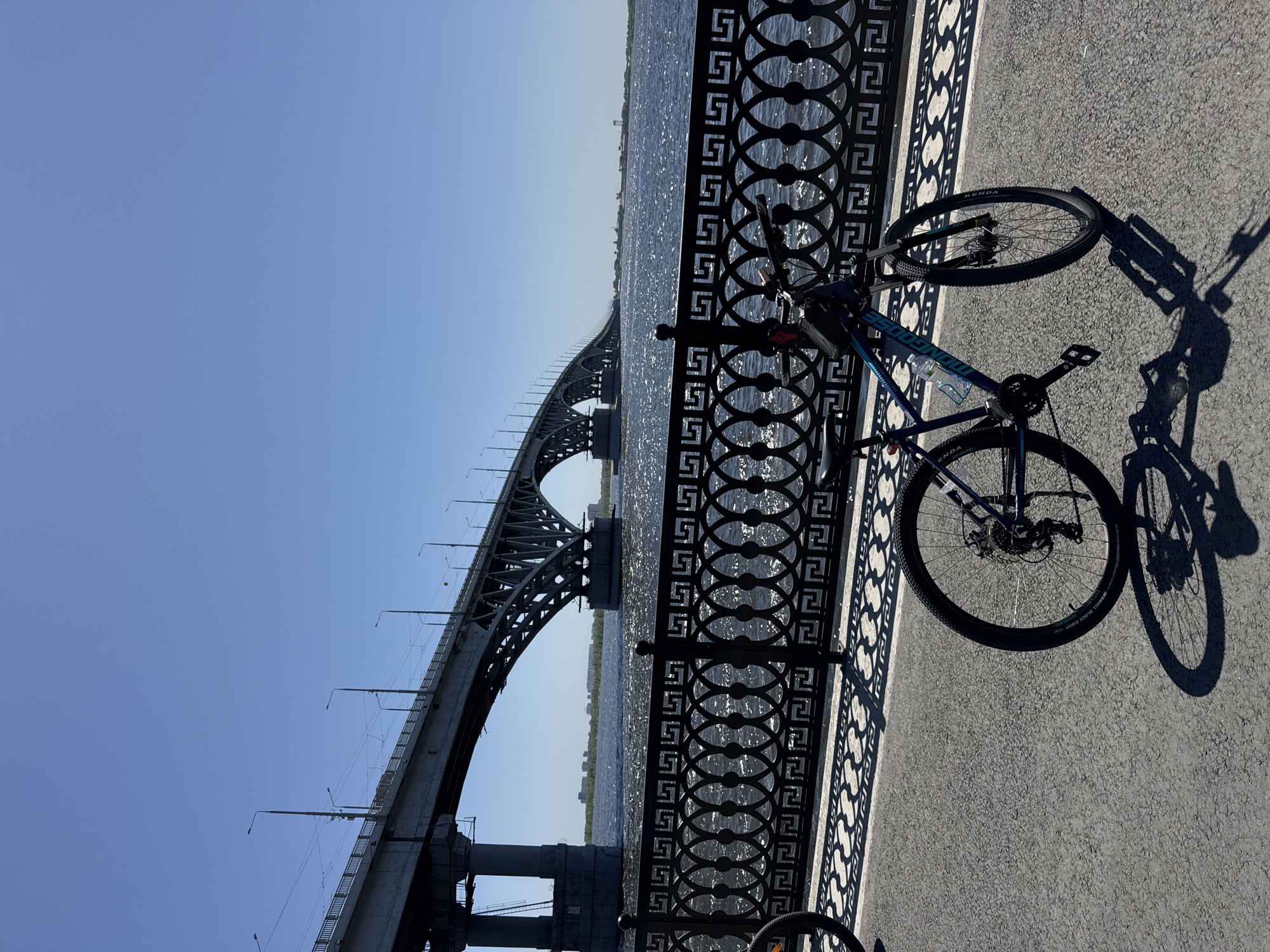 A bicycle is parked on a sidewalk next to a decorative metal railing overlooking a river. A bridge spans the river in the background. The location is likely in Saratov, Russia, given the architectural style of the bridge. The sky is clear and blue. The bike casts a shadow on the sidewalk. The river reflects sunlight.