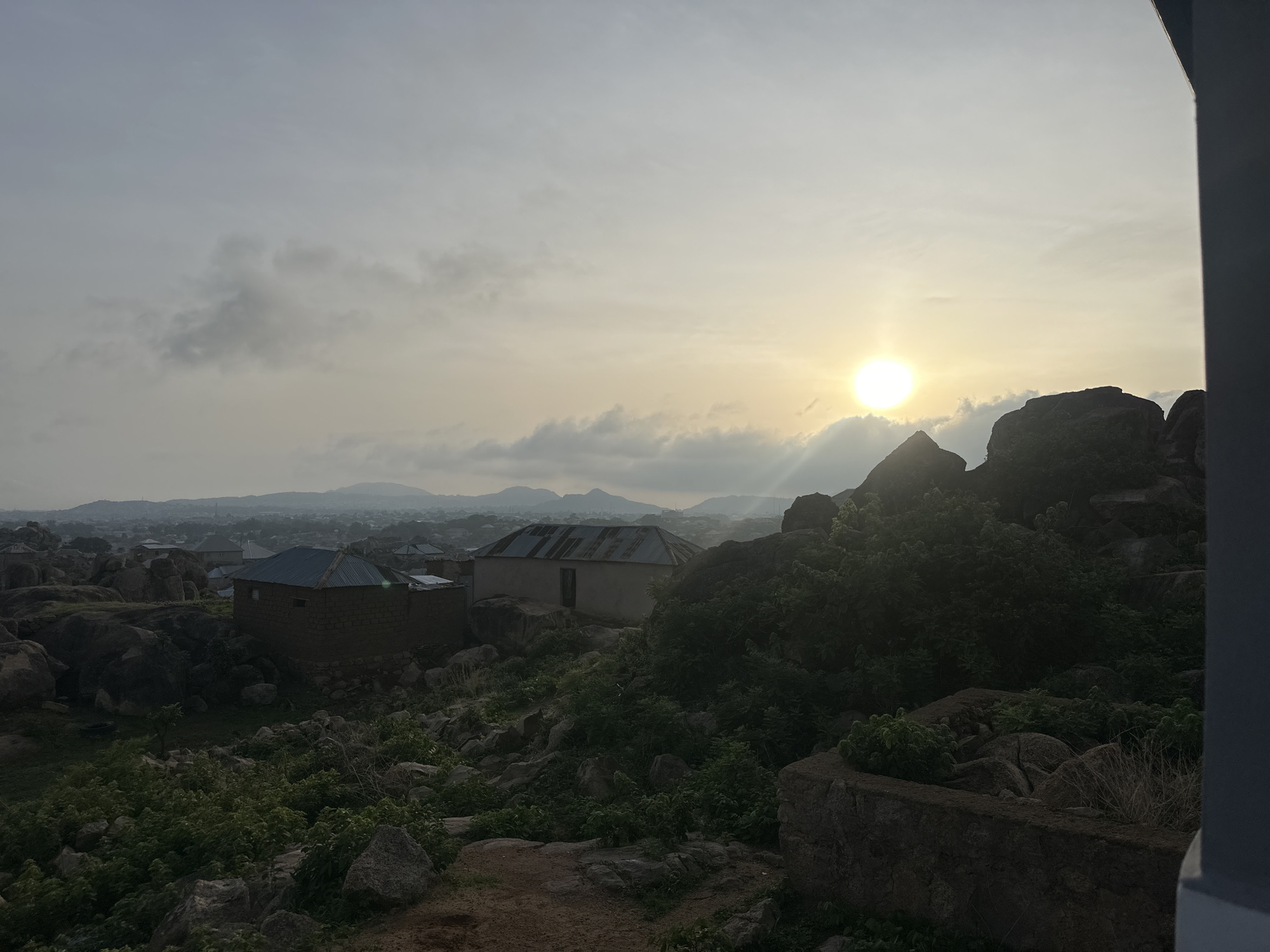 This media file presents an expansive landscape view from an elevated vantage point in what is identified as Jos, Nigeria. The scene is bathed in the soft, diffused light of either sunrise or sunset, with a prominent, bright sun partially obscured by clouds on the right side of the sky. The sky features a mix of light grey and darker clouds, contributing to an overall hazy atmosphere, especially over the distant town.
The foreground and midground are characterized by rugged, undulating terrain, dominated by numerous large boulders and natural rock formations. Green bushes and sparse vegetation grow among these rocks, adding touches of verdant color to the otherwise earthy palette. Nestled within this rocky landscape are two simple, single-story residential structures, each featuring corrugated metal roofs. One house, situated on the left, has dark-colored walls, while another, more centrally located, has lighter-toned walls, a visible door, and a small window.
Beyond the immediate rocky environment, a sprawling town or city extends into the hazy distance, with countless buildings and structures visible. A chain of hills or mountains forms the backdrop on the distant horizon, partially obscured by the atmospheric conditions. A dark, vertical architectural element partially frames the image on the right, suggesting the photograph was taken from within or adjacent to a building. No people or specific activities are visible, and there is no discernible text. The image captures a serene, natural setting with evidence of human habitation integrated into the rugged topography.
