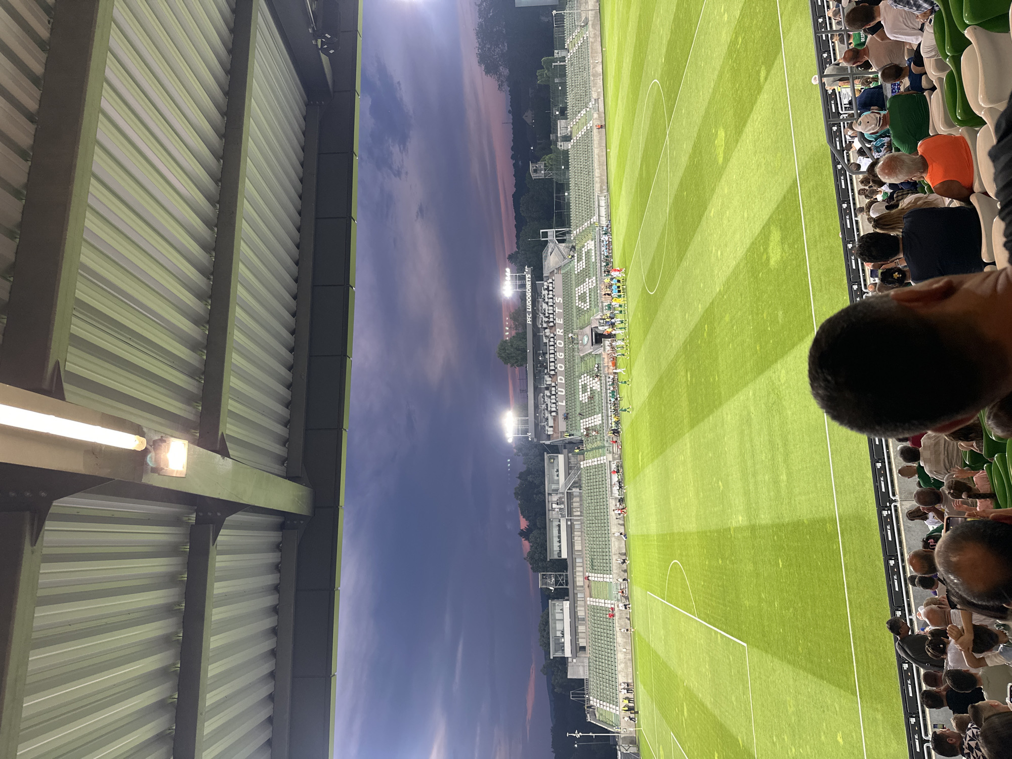 The image depicts a wide view of the Huvema Arena, home stadium of PFC Ludogorets Razgrad, in Razgrad, Bulgaria. The stadium is captured at dusk with a dramatic purple and pink sky visible above the stands and trees in the distance. The field, a vibrant green with white markings, is illuminated by stadium lights. Two teams of players, dressed in green and white uniforms, are lined up on the field, appearing to be in formation or about to start a game. Spectators are visible in the foreground, seated in green stadium seats, with some focused on the field and others looking at their phones. The architecture of the stadium, including the roof structure with corrugated metal and lighting fixtures, is also prominent in the frame. The overall scene suggests a sporting event is about to commence or has just concluded.