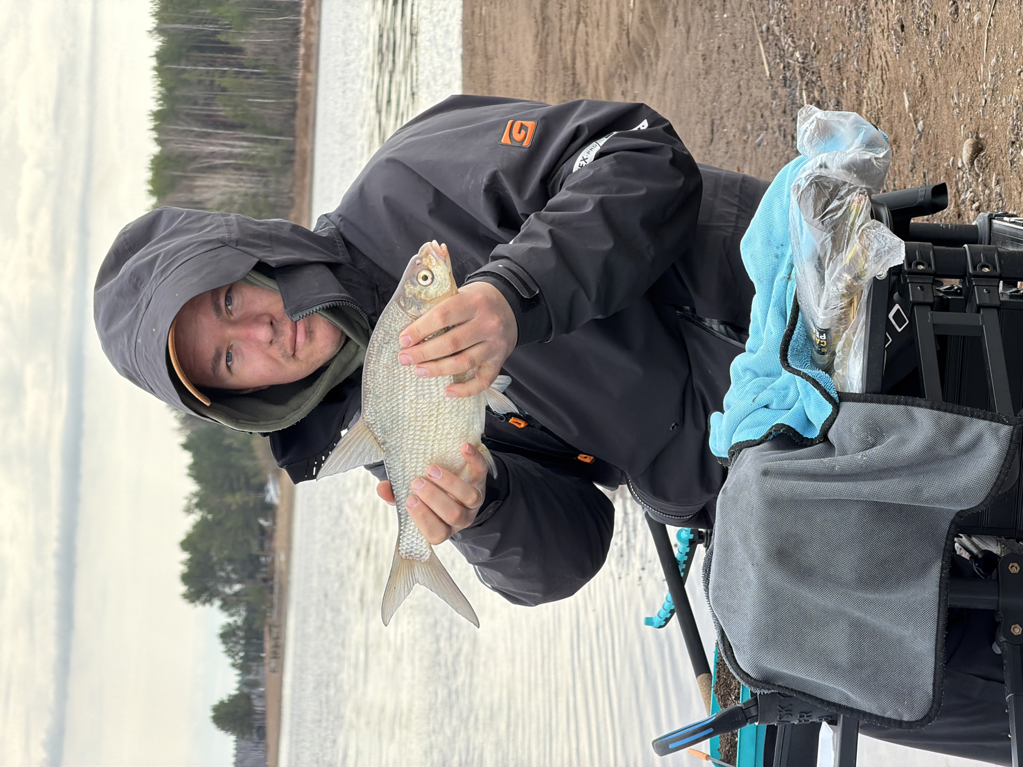 A man in a dark jacket with a hood is holding a fish. The background features a body of water, likely a river or lake, and a shoreline with trees. Other fishing equipment, like a black frame and a blue towel, are visible to the right. The scene suggests an outdoor setting, possibly near the city of Chaykovsky, Russia, given the geography.