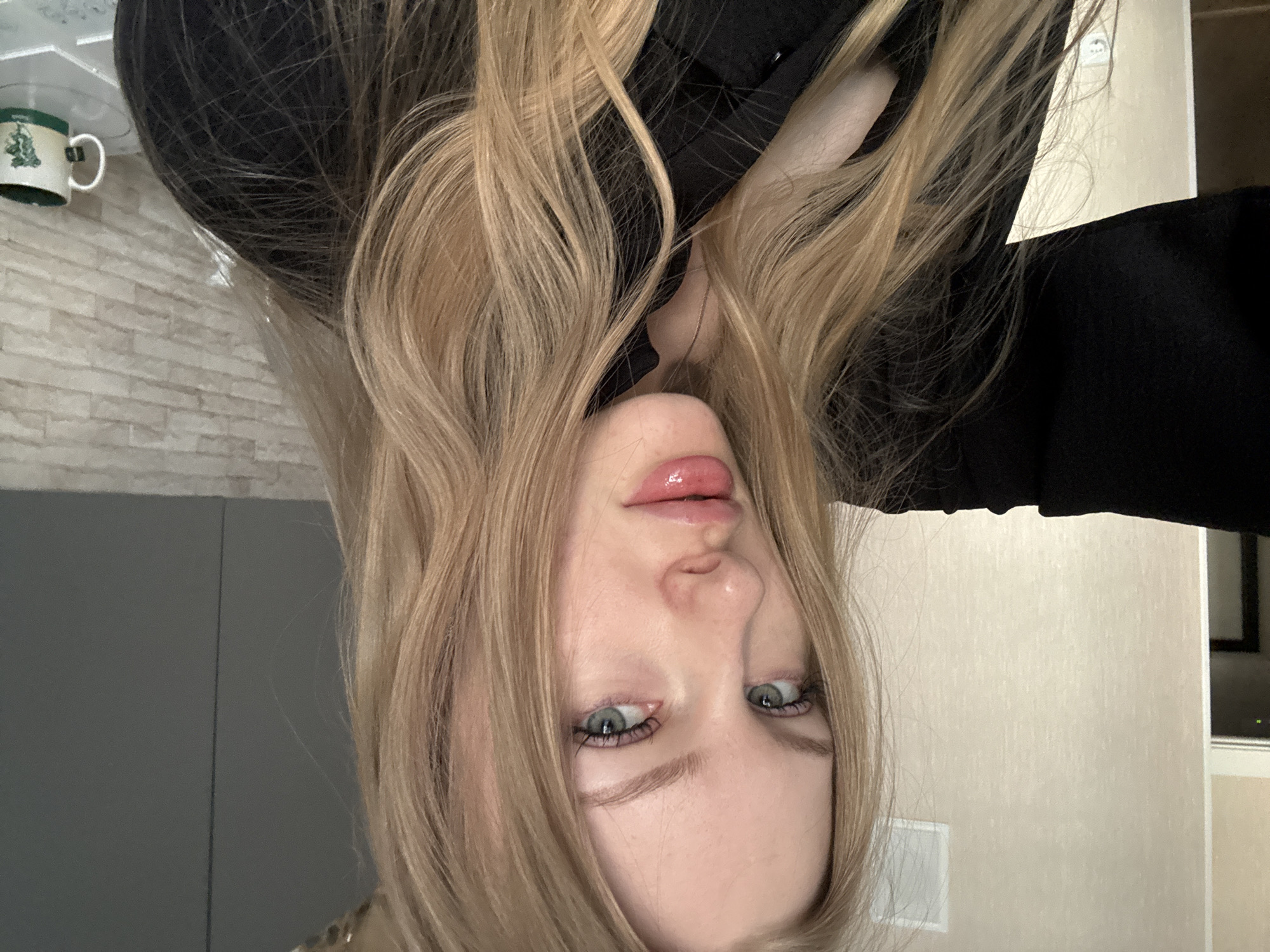 A close-up, inverted selfie shows a blonde-haired woman with fair skin and light green eyes. She is wearing a black top. In the background, a light-colored brick wall and a green mug are visible on a shelf. A gray wall is also in the background. The location is likely an interior space, possibly a residence, although specific information confirming it is in Perm, Russia, is absent.