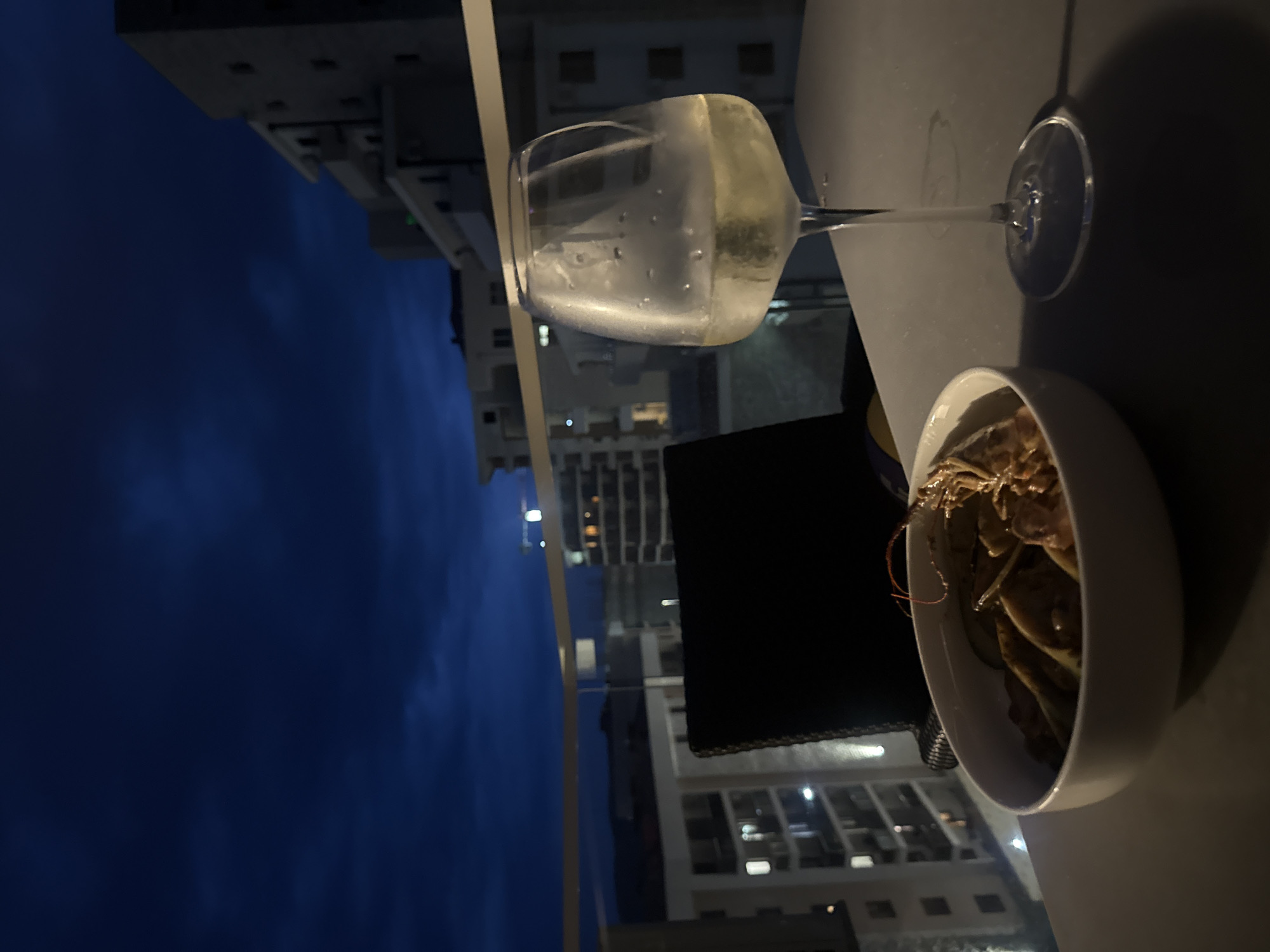 The image depicts a dining scene on a balcony at dusk, overlooking a city. In the foreground, a white bowl filled with pasta and seafood is placed on a table, to the right of which is a wine glass filled with a light-colored liquid, possibly white wine. The table casts a shadow on the surface. Behind the table, a black chair is partially visible. The background reveals a cityscape with buildings of varying heights, illuminated by artificial lights, under a dark blue sky with hints of clouds. A glass railing separates the balcony from the view. The location is identified as Boreti, Montenegro.