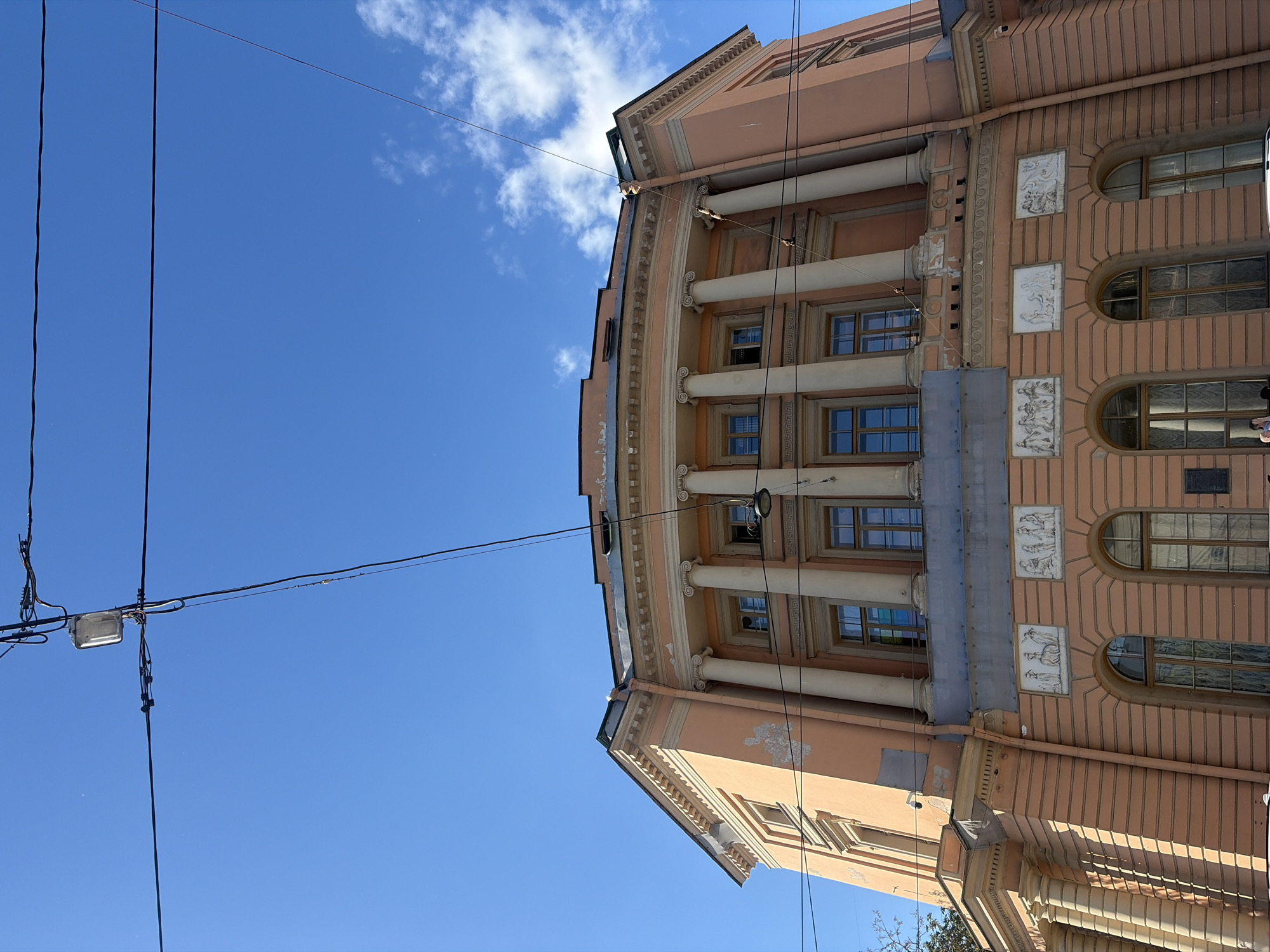 This image shows a view of the Mikhailovsky Palace in Saint Petersburg, Russia, on a clear, sunny day. The palace, painted a salmon pink, features a grand curved facade with classical architectural elements, including columns, pediments, and decorative reliefs. Several windows are visible, some with a slightly distressed appearance. Overhead, a network of electrical wires crisscrosses the bright blue sky, with a single street lamp visible. A few wispy white clouds are present in the upper right portion of the sky. The bottom of the image shows a portion of a sidewalk and what appears to be the top of a white vehicle.