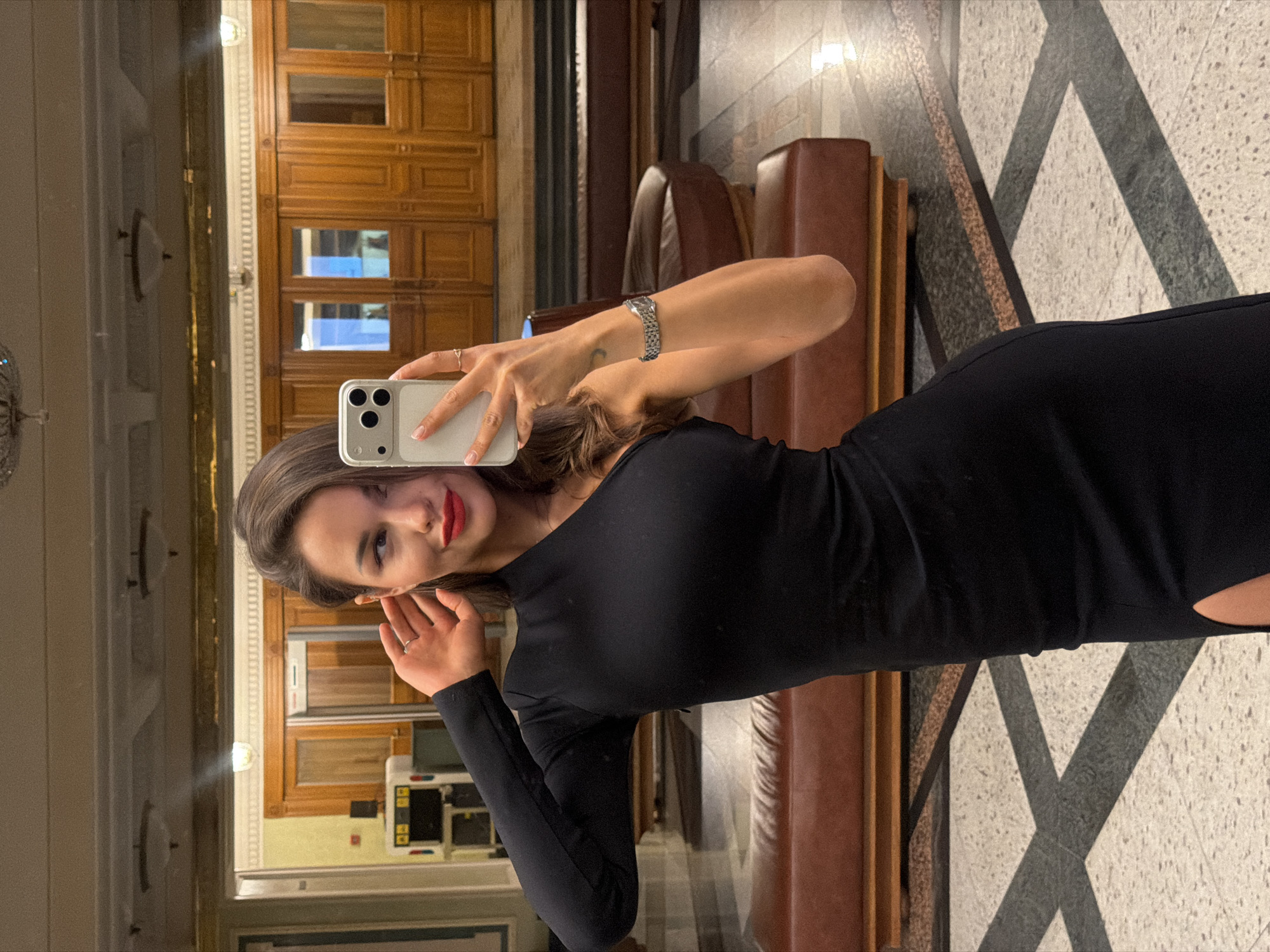 A young woman is captured in a mirror selfie, standing in what appears to be a sophisticated indoor public space, possibly a hotel lobby or a similar establishment, consistent with locations found in Moscow, Russia. She is shown from the waist up, with her body angled slightly.
The woman has medium-length brown hair, styled casually. She is wearing a fitted black long-sleeved top or dress, and her lips are adorned with red lipstick, complemented by light makeup. She has a pleasant, soft smile, looking directly at the camera in her phone's reflection. On her left wrist, she wears a silver-toned metallic link watch or bracelet, and a simple silver-colored ring is visible on her right ring finger. She holds a white or light-colored smartphone with a visible triple-lens camera array, using it to capture her reflection.
The background, visible as a reflection, features elegant architectural elements. Prominent is rich, warm-toned wooden paneling with multiple rectangular segments, suggesting traditional or classic interior design. Several brown leather upholstered seating units, likely sofas or benches, are also reflected. The floor is covered with patterned tiles, showcasing a distinctive geometric design of alternating light and dark "X" or cross-like shapes. Artificial lighting fixtures are visible on the ceiling and walls, illuminating the space, suggesting it could be evening or nighttime. No visible text is discernible in the image.