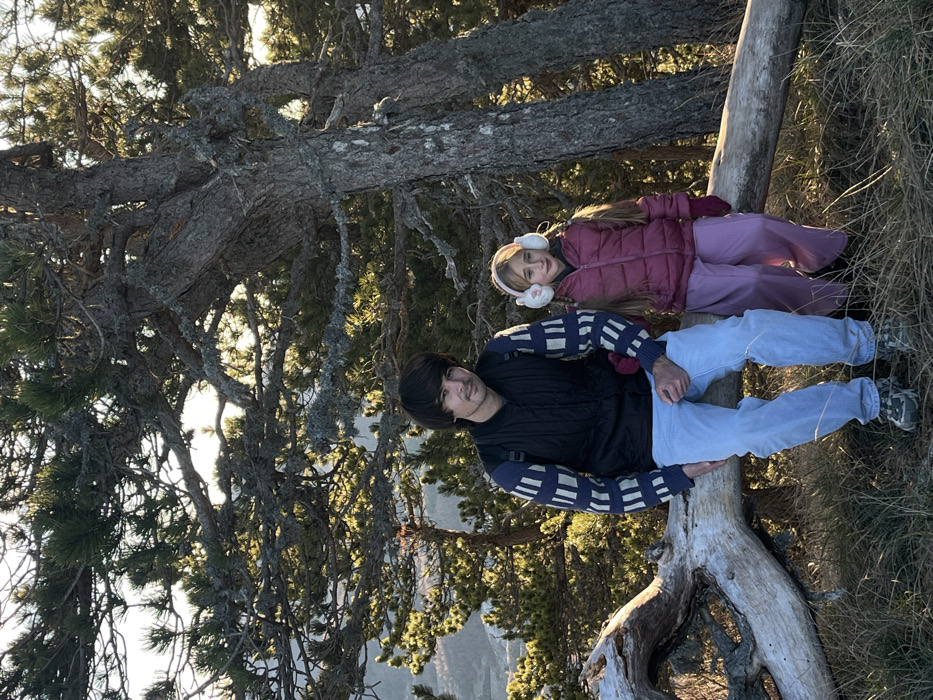 A man and a girl lie on a fallen tree trunk in a forest. The man wears a black vest over a sweater with a pattern, light blue jeans, and sneakers. The girl is dressed in a burgundy jacket, lavender pants, and white earmuffs. They are resting against the trunk, with evergreen trees and a valley visible in the background. Sunlight filters through the branches above.