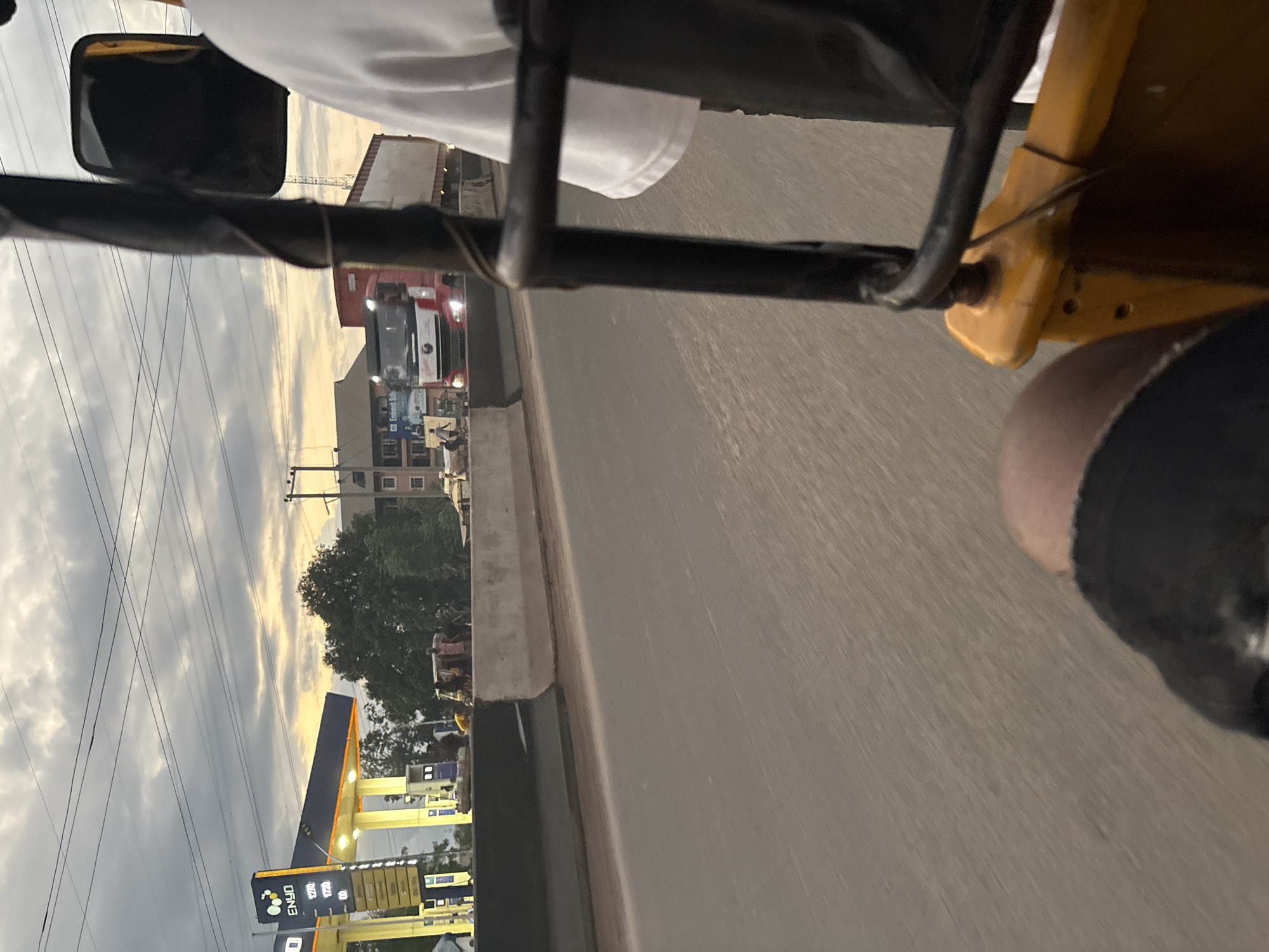 The image depicts a street scene in Onitsha, Nigeria, taken from the perspective of a passenger in a motorized tricycle (keke). In the background, a large red and white truck with a trailer is driving on the road. To the left, a brightly lit ENYO fuel station is visible with a prominent sign displaying prices. Several people are gathered near the fuel station and on the side of the road, some appearing to be interacting. The sky is overcast with hints of sunset light breaking through. The road surface is visible in the foreground, indicating movement. The overall environment suggests an urban setting, likely during dusk.