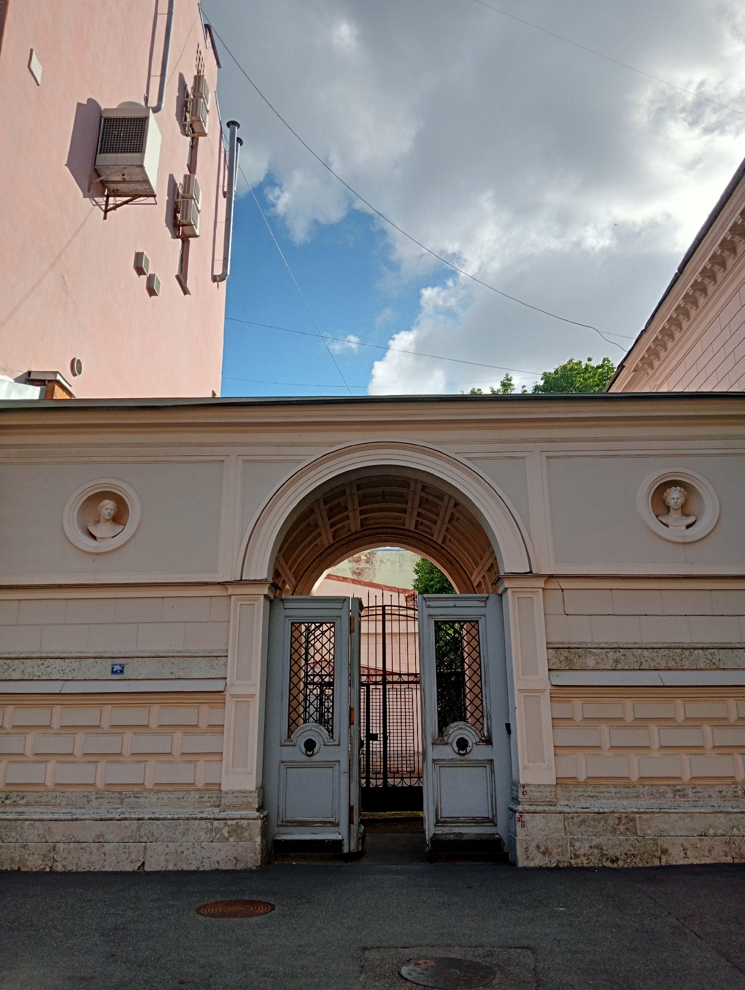The image shows a building facade in Saint Petersburg, Russia. The building is painted pink and features an arched entryway with decorative elements. The entryway leads to a wrought-iron gate. Two circular reliefs depicting busts are embedded in the wall on either side of the entryway. The sky above is partly cloudy. Air conditioning units are visible on the side of an adjacent building. The scene appears to be a street view.