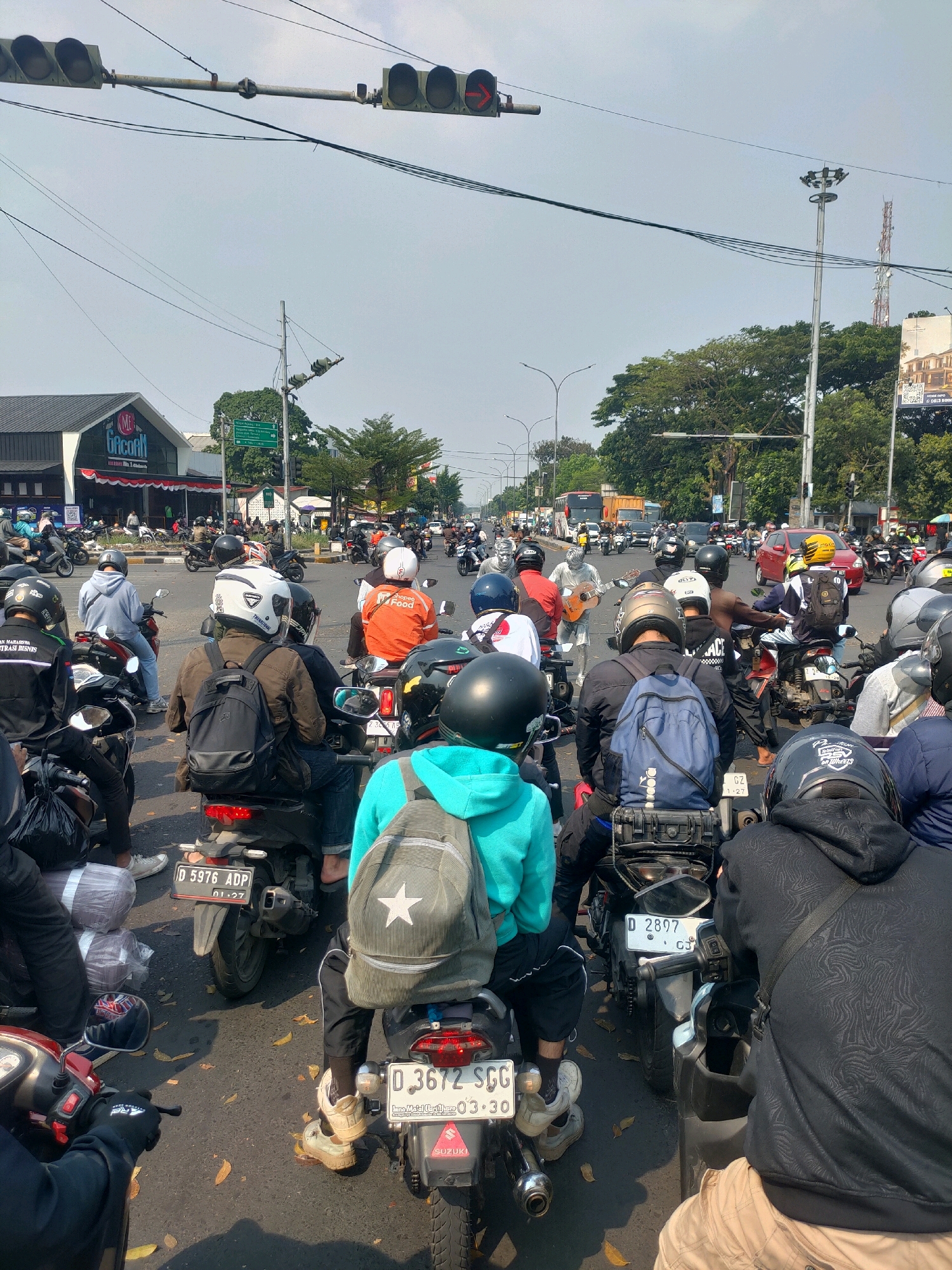 This is a street-level, medium-shot photograph taken from behind a line of motorcycles stopped at a traffic light. The scene is a busy urban intersection in Bandung City, Indonesia, suggested by the visible traffic lights, dense motorcycle traffic, and signs in Indonesian. The time of day appears to be afternoon, indicated by the bright sunlight casting distinct shadows on the asphalt.
The foreground is dominated by the backs of several motorcyclists and their vehicles. The closest subject is wearing a black hoodie with a textured pattern and beige pants. To their left, a motorcyclist in a bright teal hoodie with a white star-emblazoned backpack is visible. Behind them, a motorcyclist in a brown jacket with a black backpack is looking forward. Other motorcyclists, wearing helmets of various colors including white, black, and orange, are also visible, some with backpacks or bags. A person in a silver costume playing a guitar stands in the middle of the intersection, an unusual sight amidst the traffic.
The road surface is asphalt, scattered with fallen brown leaves. Traffic lights hang overhead, one of which shows a red arrow indicating traffic is stopped. In the background, there are buildings with signs, including one for "M. BACON," commercial establishments, trees, streetlights, and utility poles with electrical wires. Several cars and buses are also present in the traffic, contributing to the overall congestion. The sky is a pale blue with a few wispy clouds. The overall impression is one of urban mobility and a moment of pause in a bustling city environment.