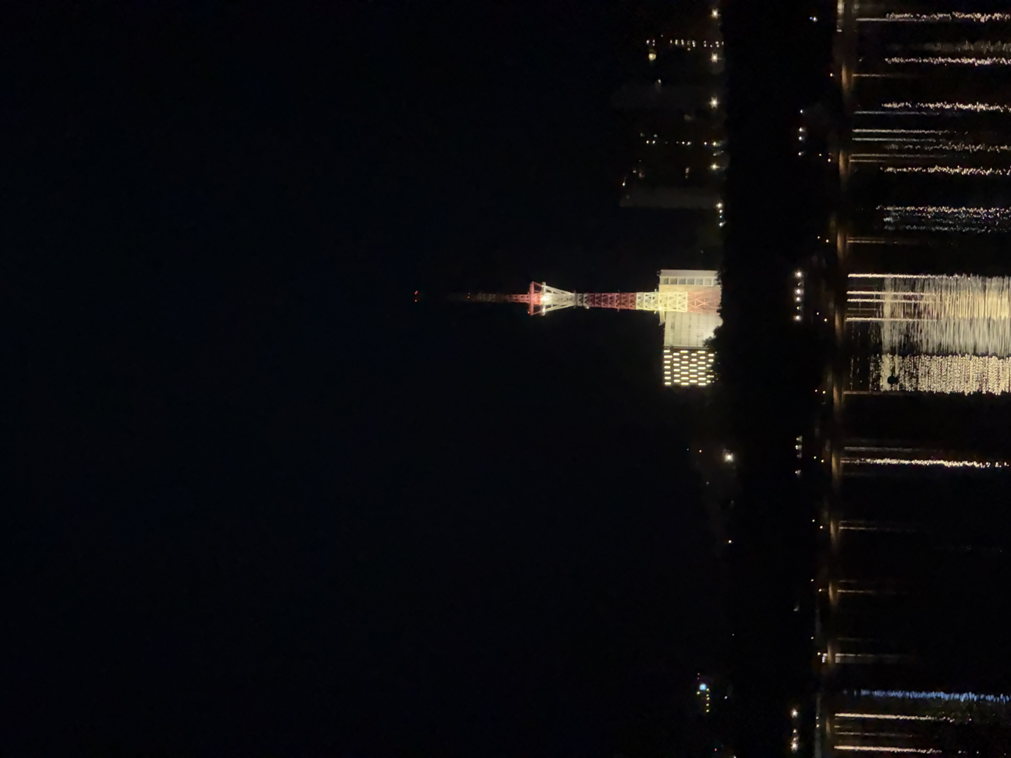 The image shows the lit-up cityscape of Perm, Russia, at night. A tower is prominently displayed, with a reddish-white structure and bright white buildings at its base. Adjacent to the buildings is a dark body of water. On the water are bright reflections of the buildings and city lights. The background is largely dark.