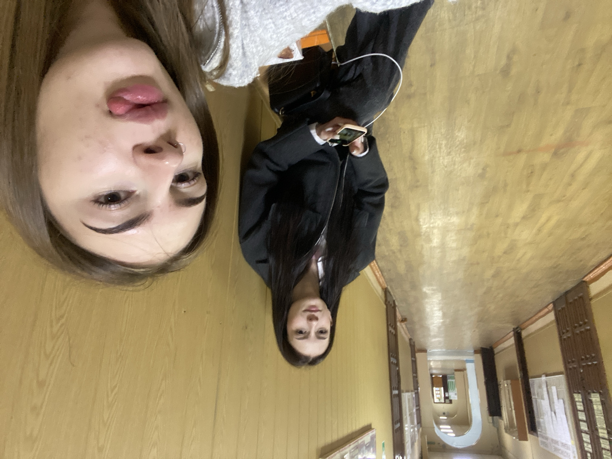 This media file captures two young women in an indoor hallway, presented with a 90-degree clockwise rotation.
In the foreground, the upper half of a woman's face is prominently displayed, appearing upside down due to the rotation. She has light brown hair, dark eyebrows, and is seen with her tongue slightly extended and lips pursed. A small silver hoop nose piercing is visible on her right nostril.
Further back and to the right, a second young woman is depicted from the waist up, also oriented sideways. She has long, dark hair and is wearing a dark jacket. She holds a light-colored smartphone in her hands, and white earphones are connected to the phone, draped over her left shoulder. She appears to be looking towards the camera.
The setting is a long, indoor hallway with light brown wooden or laminate flooring and light beige or yellow walls. The hallway extends into the background, revealing an archway and further sections of the corridor. Along the right wall, there are architectural details that resemble display boards or built-in cabinets with some papers attached. The lighting suggests an interior environment, possibly artificial. No discernible text is visible on the papers or elsewhere in the image. The overall impression is a casual, possibly playful, selfie taken in a public or institutional building like a school or office.
