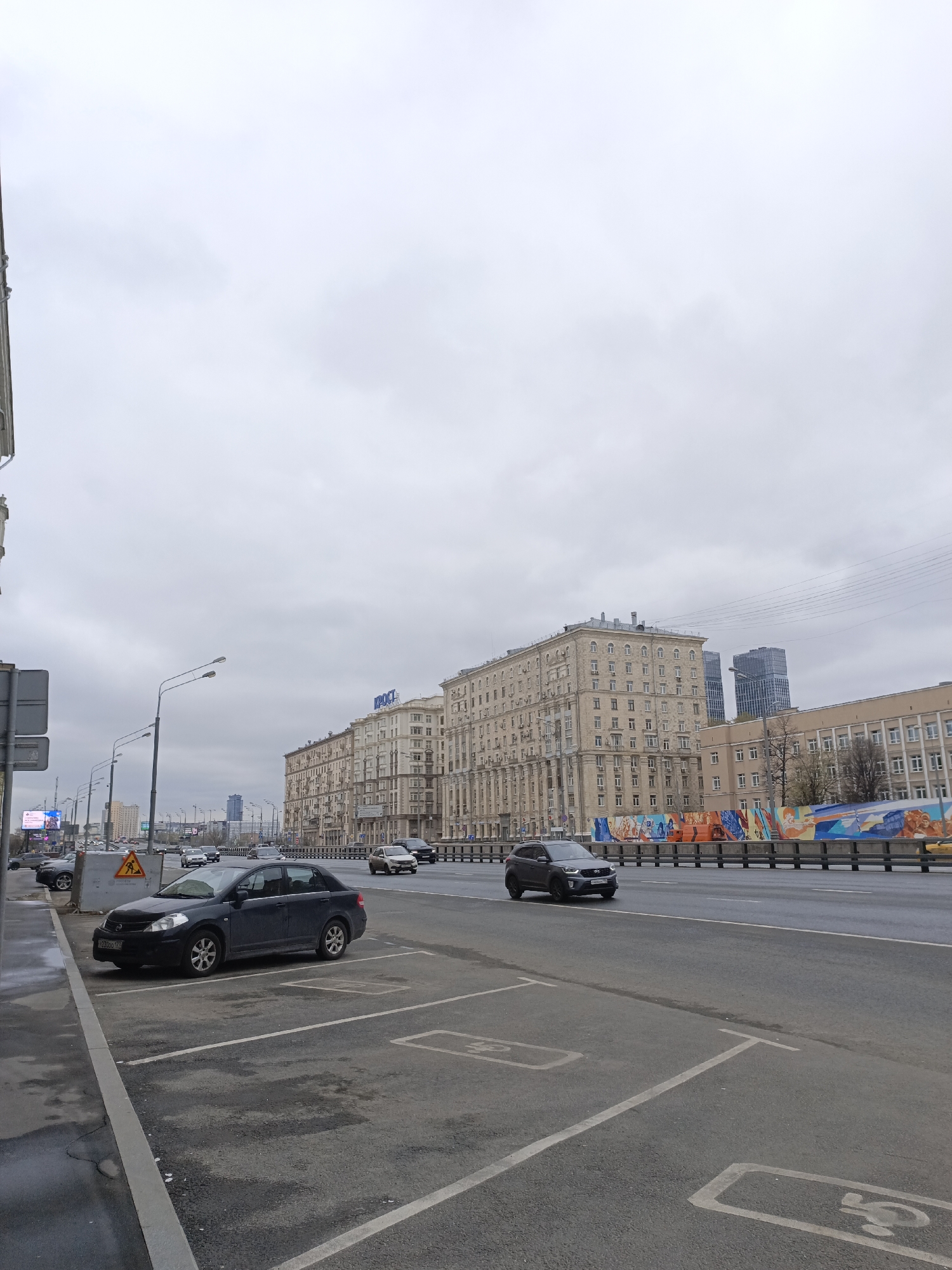 The image depicts a street scene in Moscow, Russia, on an overcast day. A dark blue Nissan Tiida is parked in a designated parking space. In the background, a row of buildings is visible, with the word "KPOCT" (presumably "KROST" in Cyrillic) on top of one of them. A modern skyscraper can be seen in the distance behind the buildings. A concrete overpass is decorated with a colorful mural. Several cars are on the road, driving in both directions. The parking spaces are marked with symbols, suggesting they might be reserved for disabled individuals. The road surface appears wet, likely from recent rain.