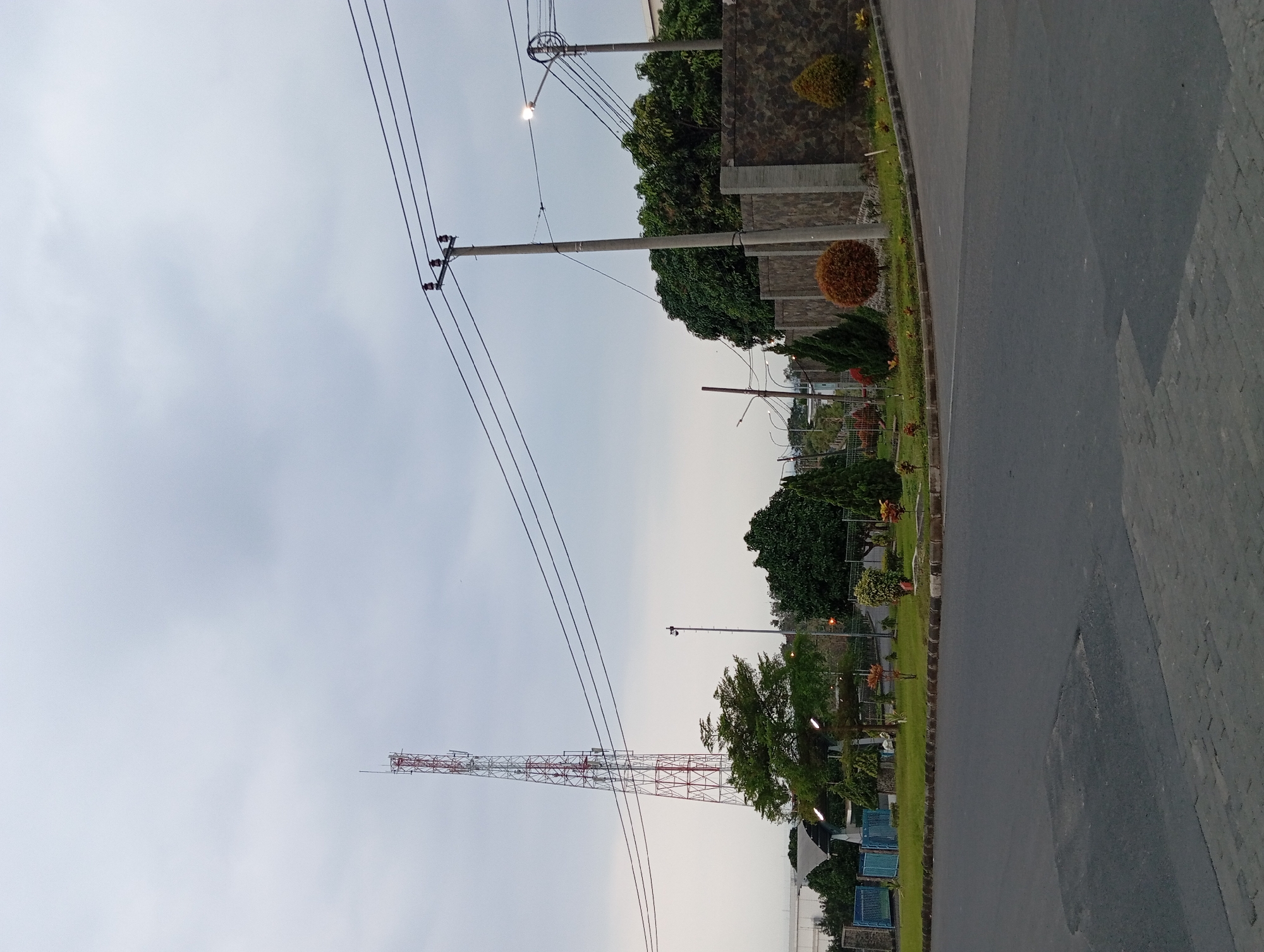 A telecommunications tower stands tall on the left of the image, its red and white lattice structure piercing the overcast sky. In front of it, a row of neatly trimmed green bushes and small trees lines a grassy area. Beyond the foliage, a light blue metal fence encloses a building with a curved roof structure.
In the mid-ground, a stone wall, possibly part of a property boundary, stretches across the scene. A tall utility pole with multiple wires extending from it stands to the right of the tower, accompanied by another shorter pole with a light fixture. Several trees with dense green foliage are visible behind the stone wall. A small, round tree with reddish-brown foliage is positioned in front of the wall, with other ornamental plants scattered around.
The foreground is dominated by a grey asphalt road with some patchwork repairs. The sky is a uniform light grey, indicating a cloudy day. The overall scene depicts an industrial or commercial area in Sumber Suko, Indonesia, with a mix of infrastructure, landscaping, and a large telecommunications tower. There are no people visible in the image.