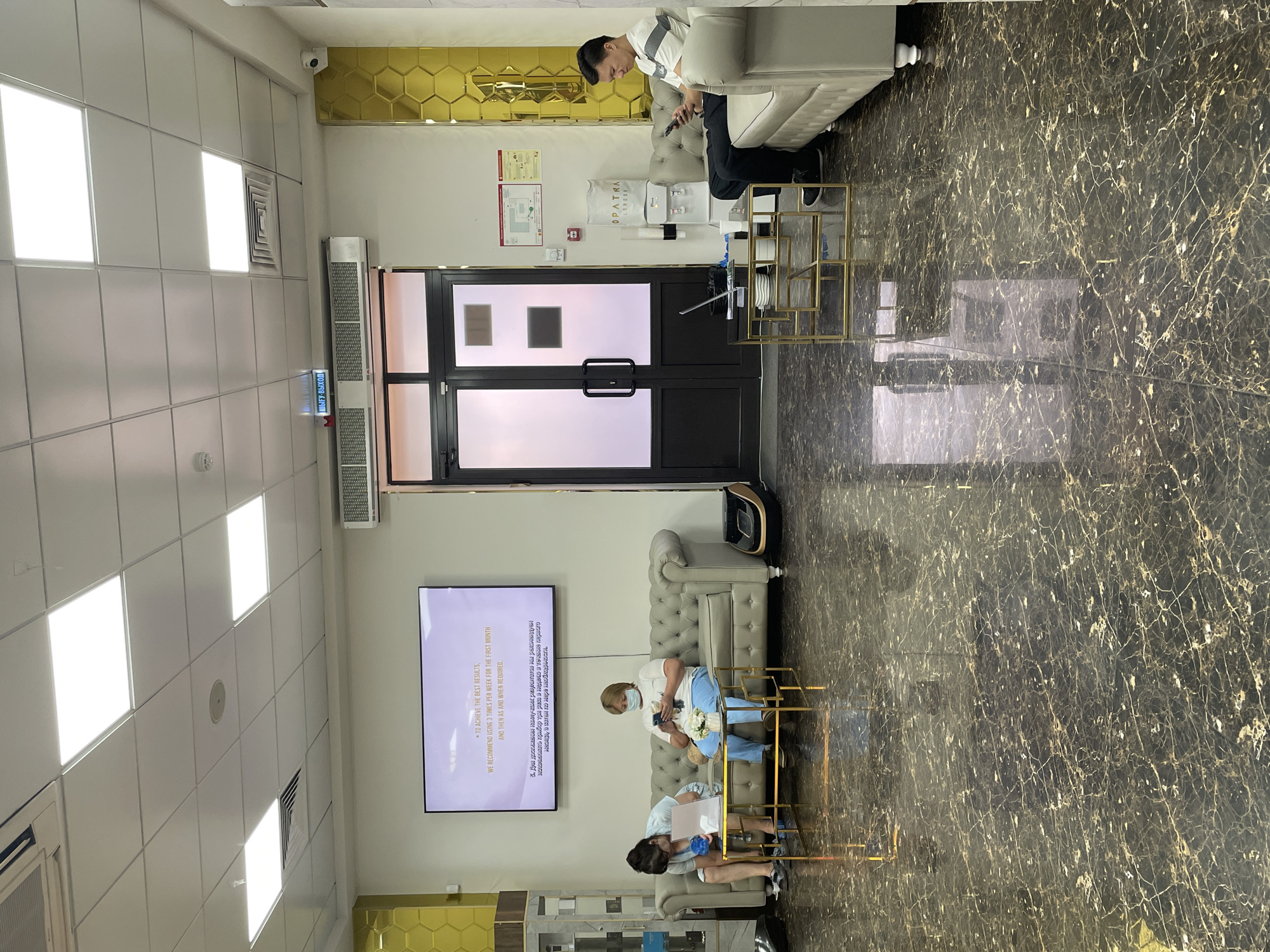 A modern, well-lit waiting area appears to be in a beauty salon or clinic in Almaty, Kazakhstan. The space features a dark, reflective marble-patterned floor, a light grey tufted sofa with two women seated, and another light grey sofa with a man seated on the right. In the center, a man and woman are sitting on the sofa, each looking at their mobile phones. To the left, another woman, wearing blue gloves, is seated and appears to be holding a small object or tissue. In front of the sofas are modern gold-colored coffee tables with laptops, cups, and other items. The walls are light-colored with decorative gold hexagonal panels on the right and a large screen displaying text on the left. A set of frosted glass double doors with black frames is visible in the background, with an illuminated "EXIT" sign above them. A water cooler with the brand "OPATRA LONDON" is situated near the doors. The overall ambiance is clean, contemporary, and professional.
