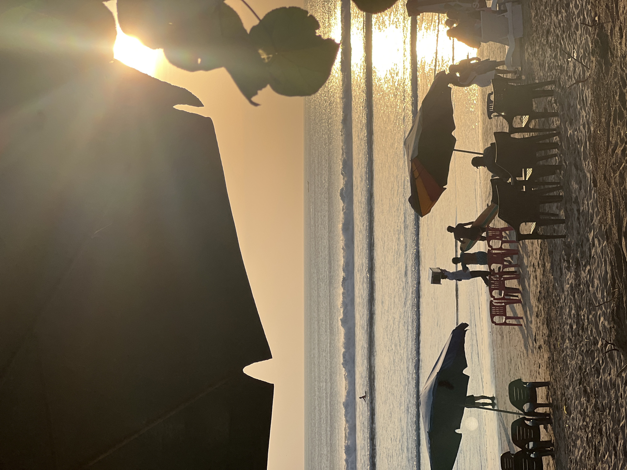 An overhead shot captures a tranquil beach scene in Kuta, Indonesia, bathed in the warm, golden light of a setting sun. The sky is a soft gradient of orange and yellow, with the sun peeking through a large umbrella in the foreground, casting lens flares and illuminating the water with a shimmering reflection. Several beach umbrellas are strategically placed on the sandy shore, some with chairs arranged beneath them, suggesting a relaxed, leisurely atmosphere. A few people are visible on the beach, including a solitary surfer in the distance, appearing as a small silhouette against the vast ocean. The gentle waves of the ocean create a calming rhythm, with the horizon marked by a distant boat. The foreground is dominated by the dark silhouette of a large umbrella, framing the sunlit seascape and adding depth to the composition. The overall mood is one of serene beauty and peaceful ambiance.