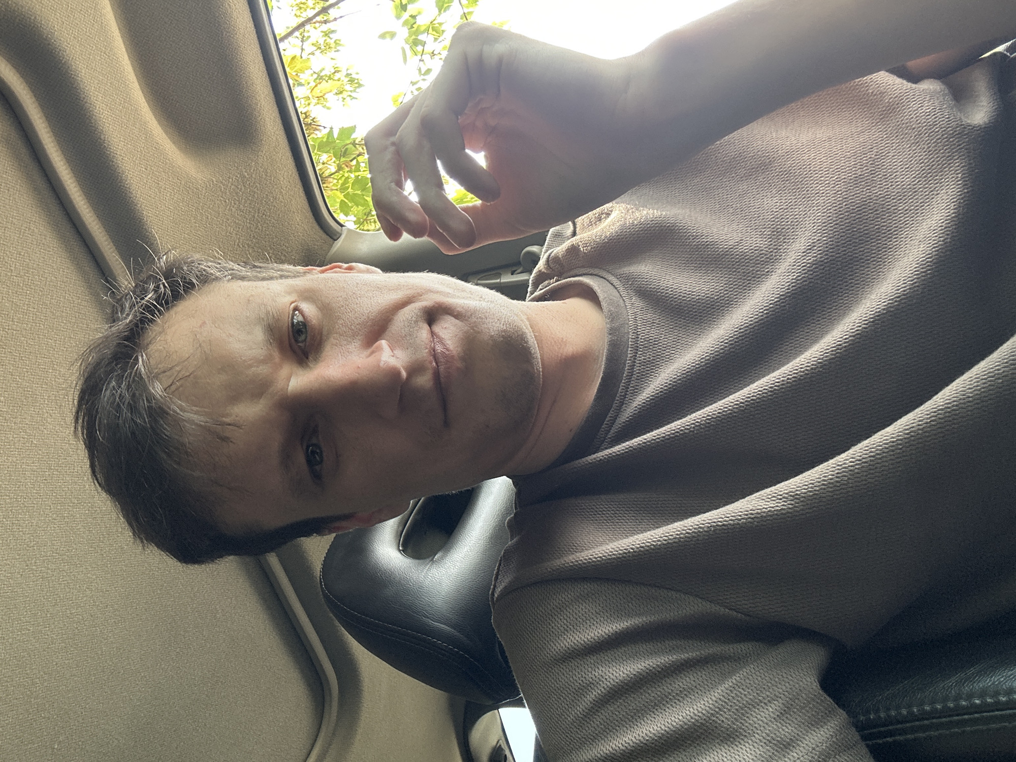 The image is a close-up, eye-level, selfie-style photograph of a fair-skinned man in his late 30s or early 40s, taken inside a vehicle. He has short, messy, light brown hair with some graying at the temples. His face is partially illuminated by sunlight coming from the right. He is looking directly at the camera with a neutral expression, his mouth slightly downturned. His right hand is raised near his face, with his thumb and index finger forming a circle. He is wearing a taupe-colored t-shirt with a textured weave. The interior of the vehicle is visible, including the textured headliner of the sunroof and the dark leather of the headrest of the seat behind him. Outside the car window, to the right, green foliage and branches of a tree are visible, suggesting an outdoor setting, potentially in a rural area. The image does not contain information that would allow for its specific location within the city of Makiv, Ukraine.