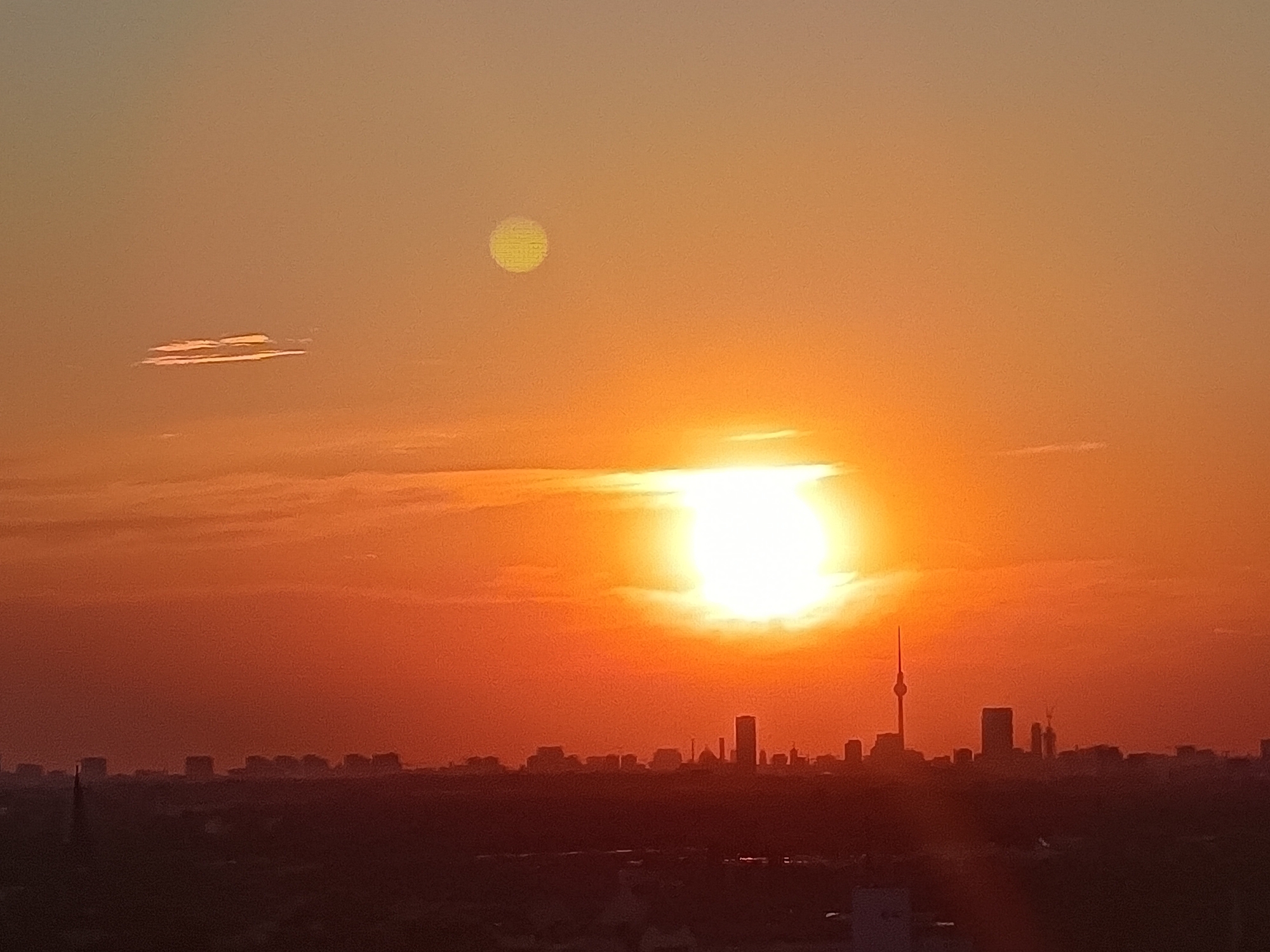The image depicts a sunset over Berlin, Germany. The sky is a gradient of orange and yellow, with the sun prominently positioned slightly to the right of the center, casting a bright glow. A smaller, pale yellow orb, likely the moon or a sun spot artifact, is visible in the upper left quadrant of the sky. A thin, horizontal cloud formation is present in the upper left, and a more diffuse band of clouds stretches across the horizon. Silhouetted against the vibrant sky is the Berlin cityscape, featuring various buildings and the iconic Fernsehturm (television tower) to the right of center. The foreground shows a dark, indistinct urban landscape. There are no people or animals visible in the frame. The overall impression is a tranquil urban sunset.